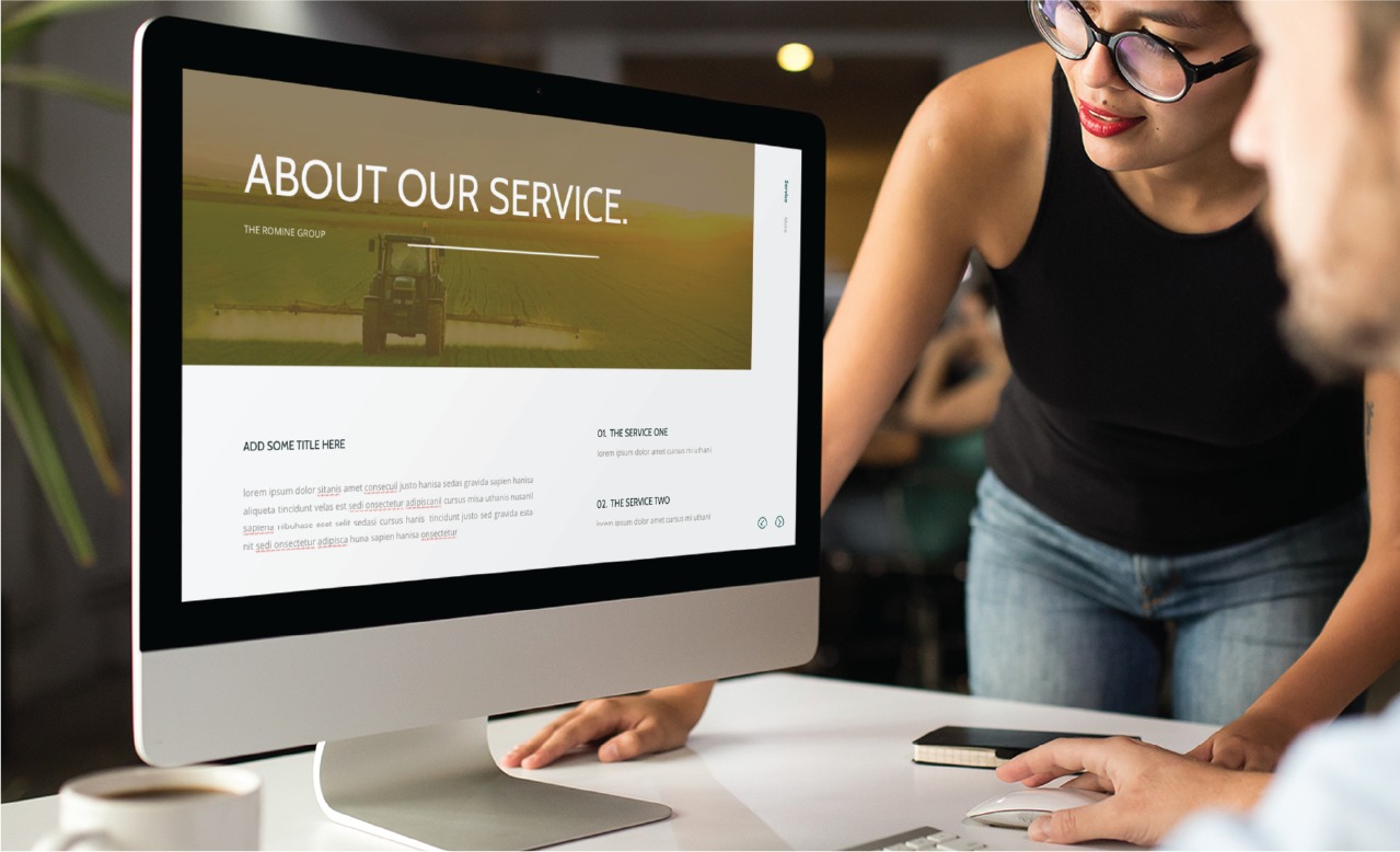 Two colleagues review a presentation on a large desktop monitor showing a slide titled'About Our Service'.