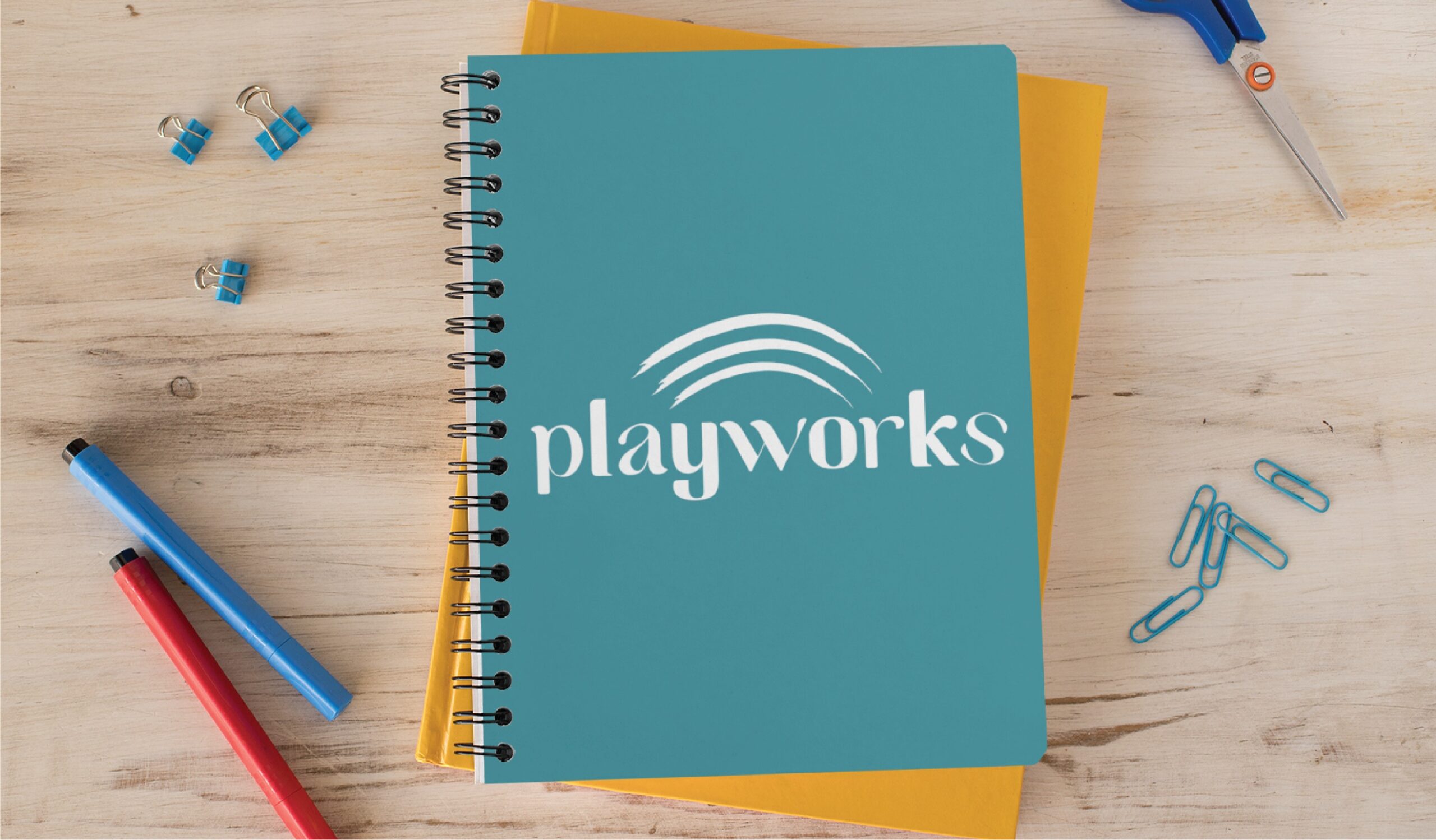 Teal spiral notebook with the Playworks logo on a wooden desk, stacked over a yellow folder, surrounded by blue pens, paper clips, and scissors.