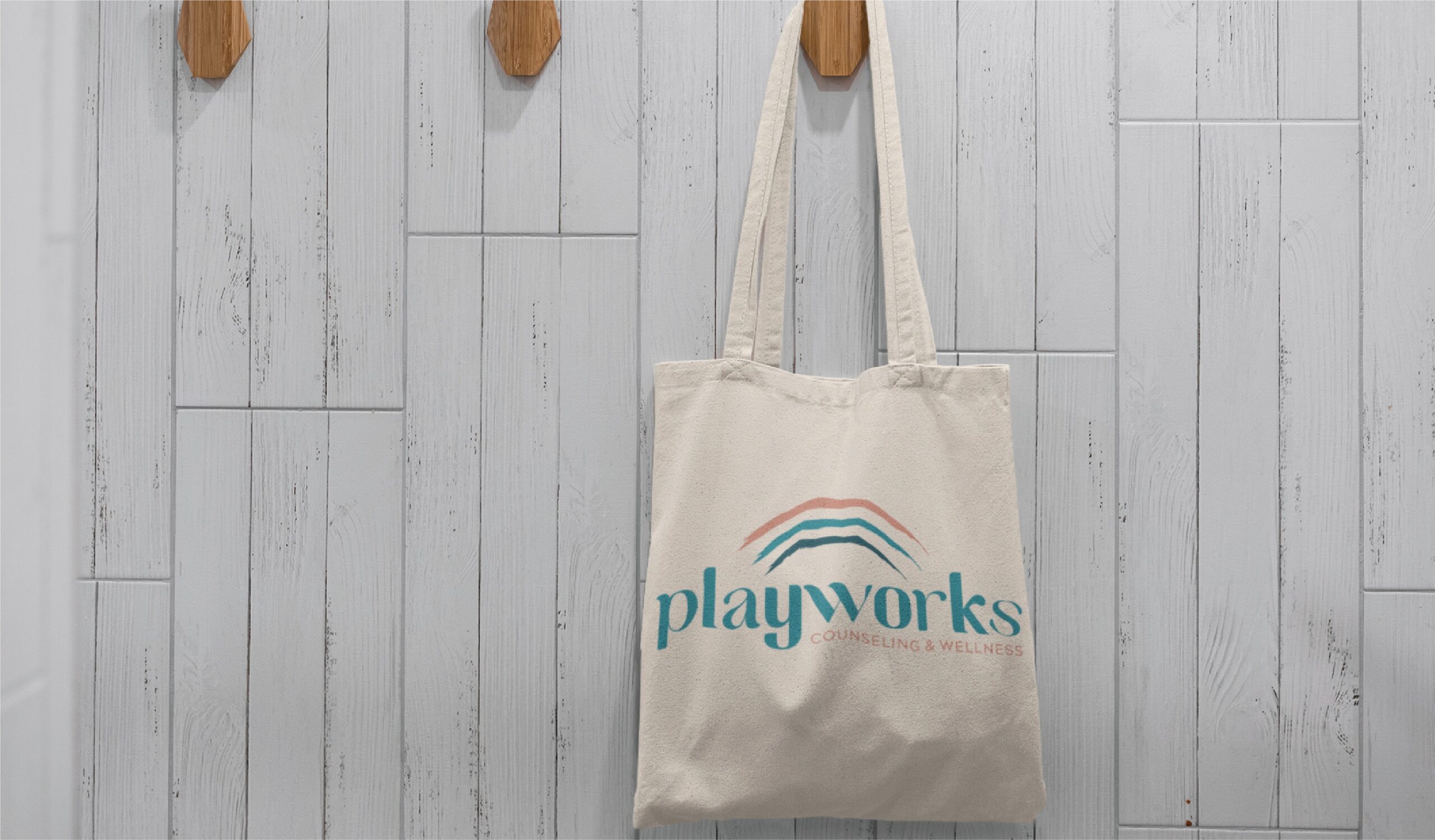 A beige tote bag with a custom logo design reading “playworks COUNSELING & WELLNESS” and a colorful arc logo, ideal for a children’s therapist in Virginia, hangs on a wooden hook against a light gray tiled wall.
