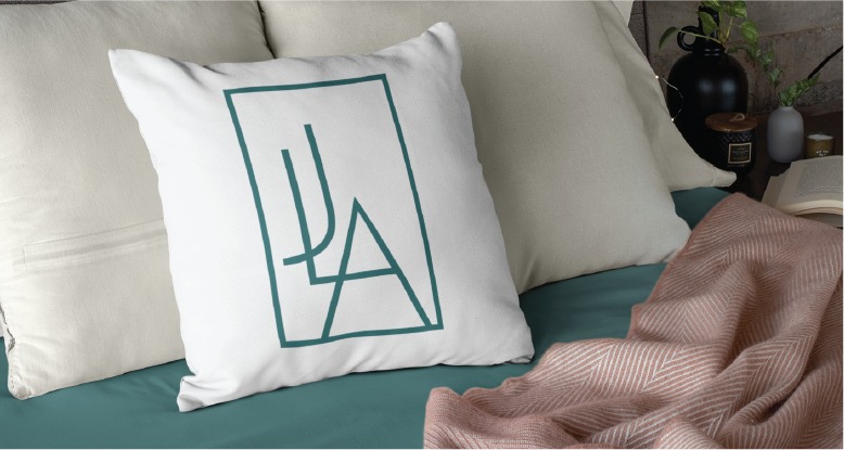 A white decorative pillow with a minimalist green geometric design rests on a bed with cream pillows, a teal sheet, a pink patterned blanket, a black lamp, and an open book nearby—creating a cozy scene that would delight any Virginia children's therapist looking for logo design inspiration.