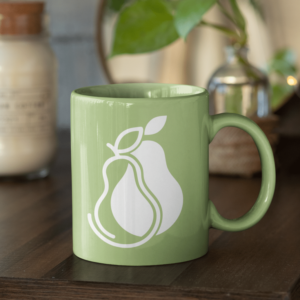 A green ceramic mug with a white outline of two overlapping pears—a nod to logo creation—sits on a wooden surface, with a candle and a plant blurred in the background.