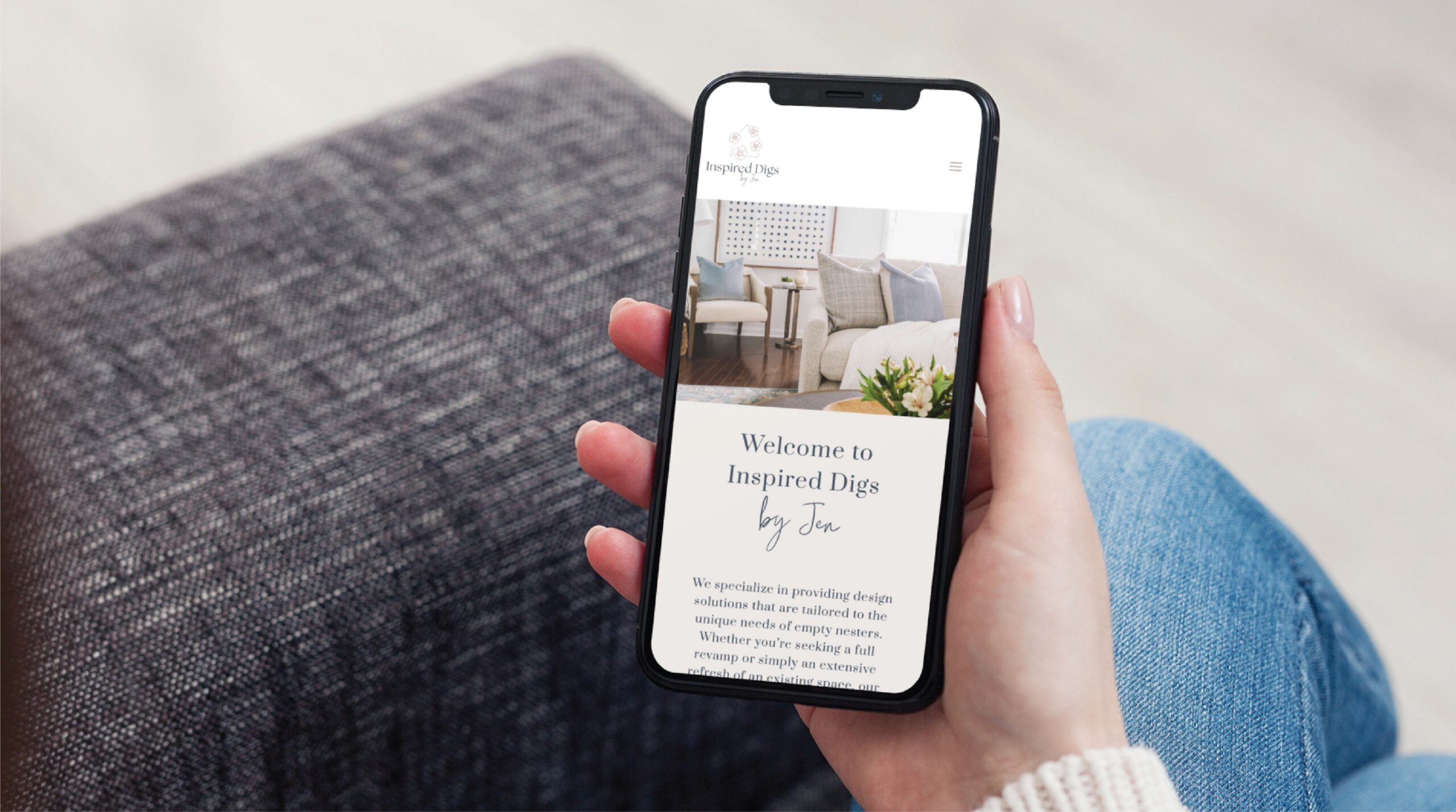 A person sitting on a gray couch in Texas, wearing jeans, holds a smartphone displaying the Inspired Digs by Jen home decor website. The cozy, decorated living room and welcome message highlight standout web design skills.