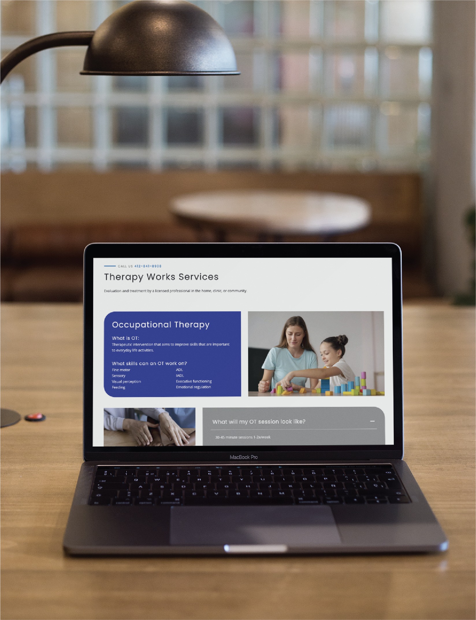 Laptop on a wooden desk showing a Therapy Works Services webpage on screen, with a lamp overhead and a blurred classroom backdrop.