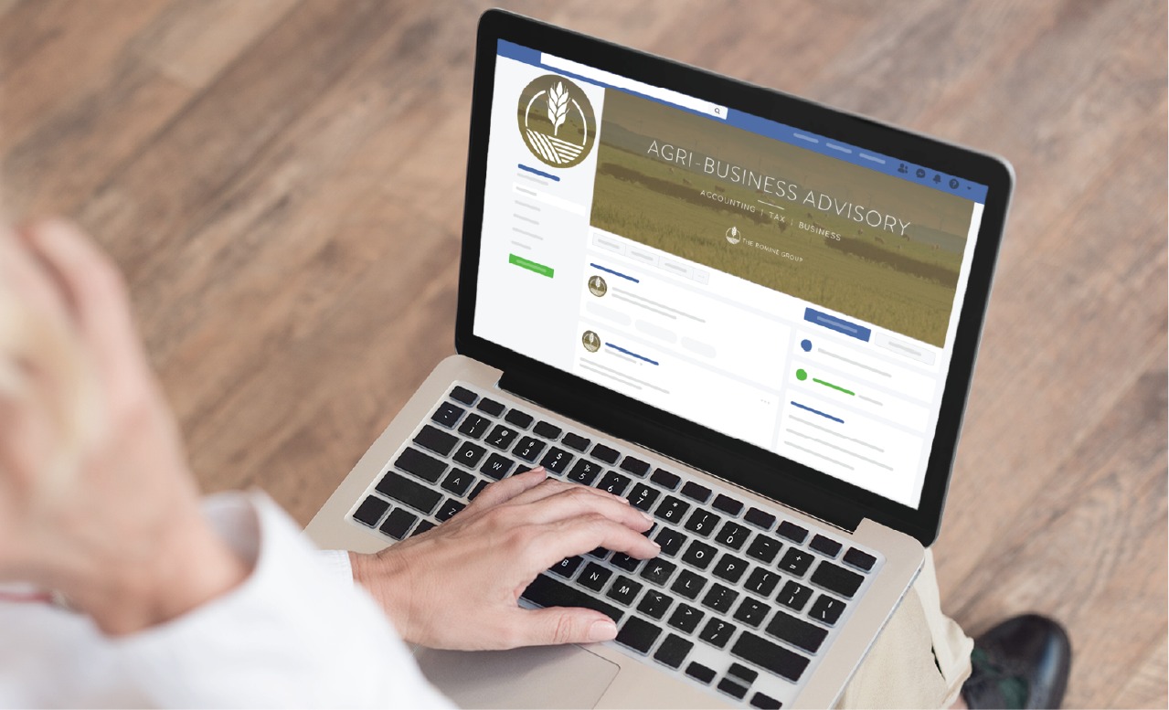 A person uses a laptop displaying a Facebook page for Agri-Business Advisory, featuring a gold and green banner with a wheat logo—showcasing thoughtful social media design. The hand is on the keyboard, and the wooden floor is visible.