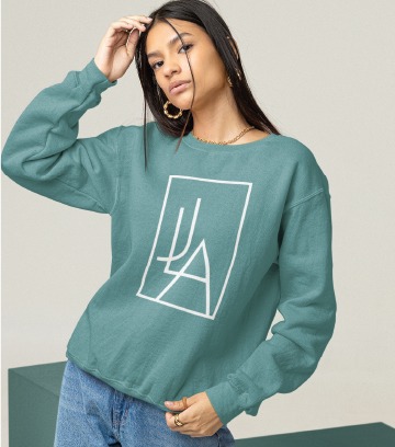 A woman wearing a teal sweatshirt with a white geometric UA logo design poses with one hand touching her head and the other at her waist, standing against a plain background.