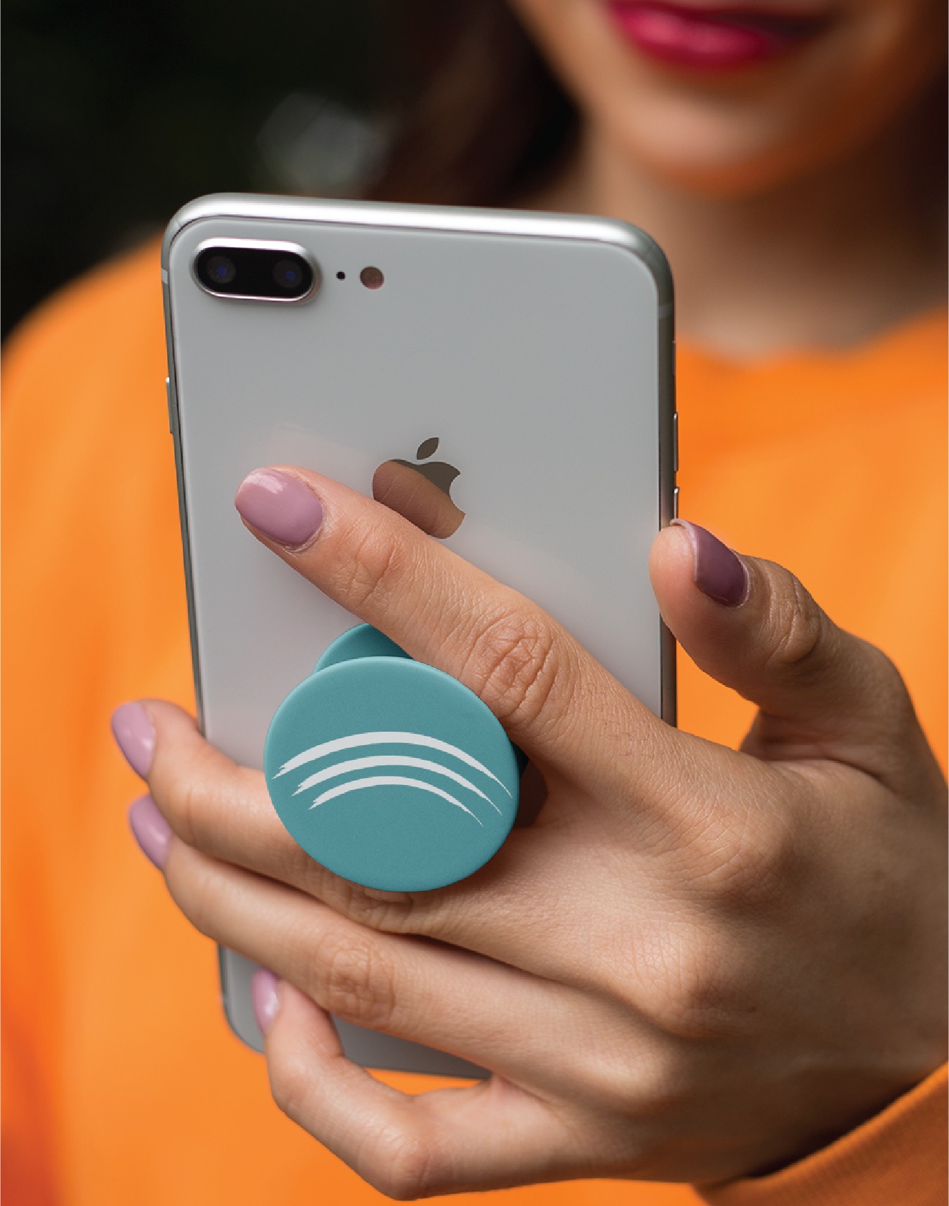A person with painted nails holds an iPhone with a teal pop socket. The person is wearing an orange top, and only the lower part of their face is visible, showing pink lipstick. The stylish setup reflects a flair for custom logo design, perfect for a creative Virginia entrepreneur capturing inspiration on the go.