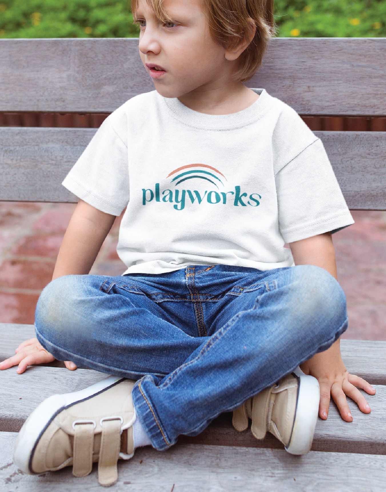 Child sitting on a bench wearing a white t-shirt with a teal'playworks' logo and arc design, jeans, and sneakers in an outdoor setting.