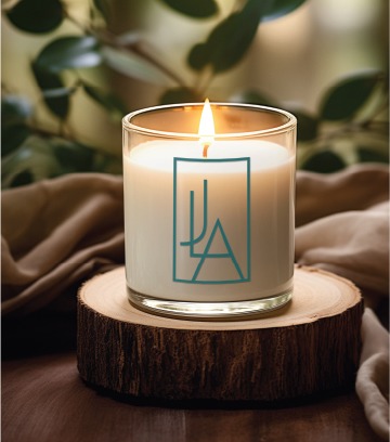 A lit white candle in a glass holder sits on a round wooden slab, with a blue geometric logo on the glass inspired by thoughtful logo design. Soft fabric and green leaves are blurred in the background, evoking the calming atmosphere of a Virginia children’s therapist’s office.