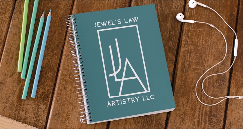 A teal spiral notebook labeled Jewels Law Artistry LLC, representing a talented Jewelry Artist in Virginia, lies on a wooden table next to three colored pencils and white earbuds.