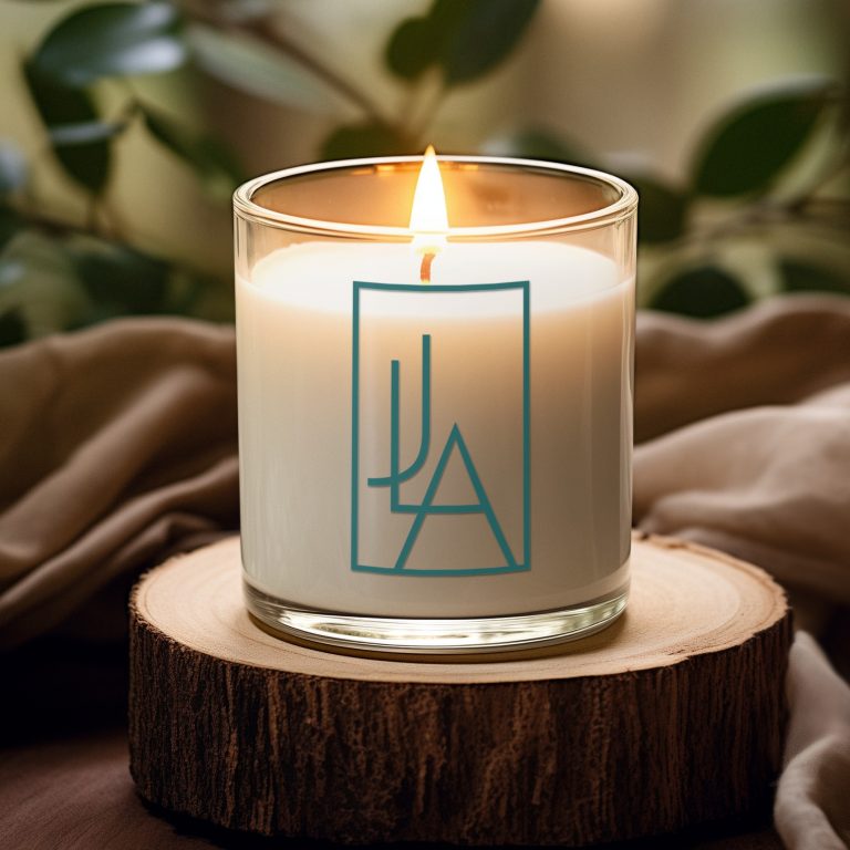 A lit white candle in a glass jar with a custom logo design in blue geometric style sits on a wooden coaster, surrounded by beige fabric and green leaves in the background.