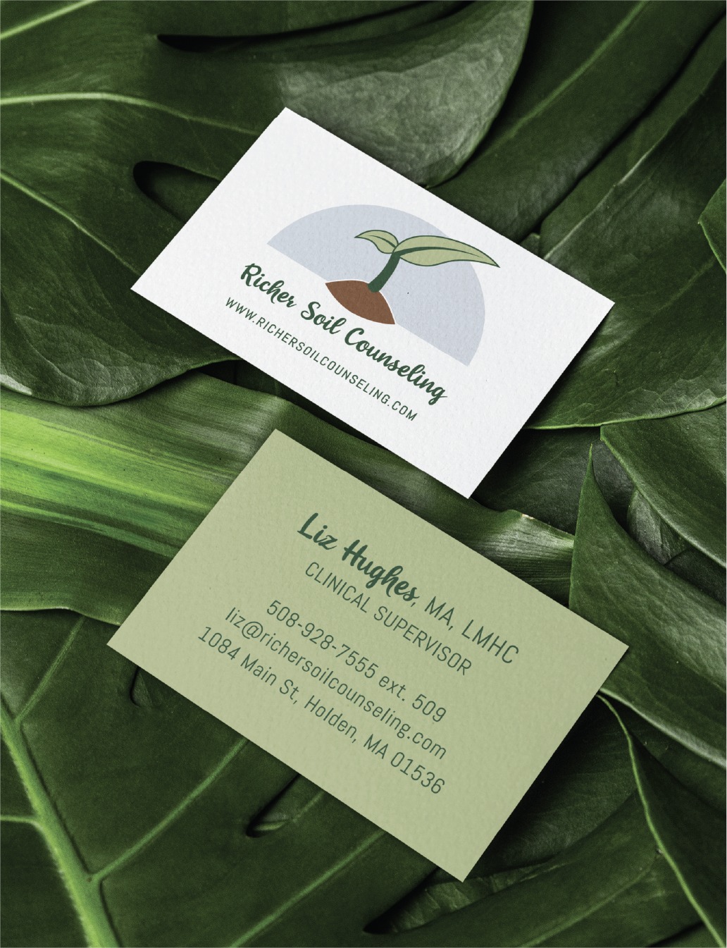 Two business cards on large green leaves showcase striking stationery design. One card features a plant seedling logo and the website richersoilcounseling.com, hinting at growth and renewal. The other card lists Liz Hughes’ contact info and the Richer Soil Counseling address in Holden, MA. These cards make an impression whether you’re seeking counseling locally or sports performance coaching as far away as California.