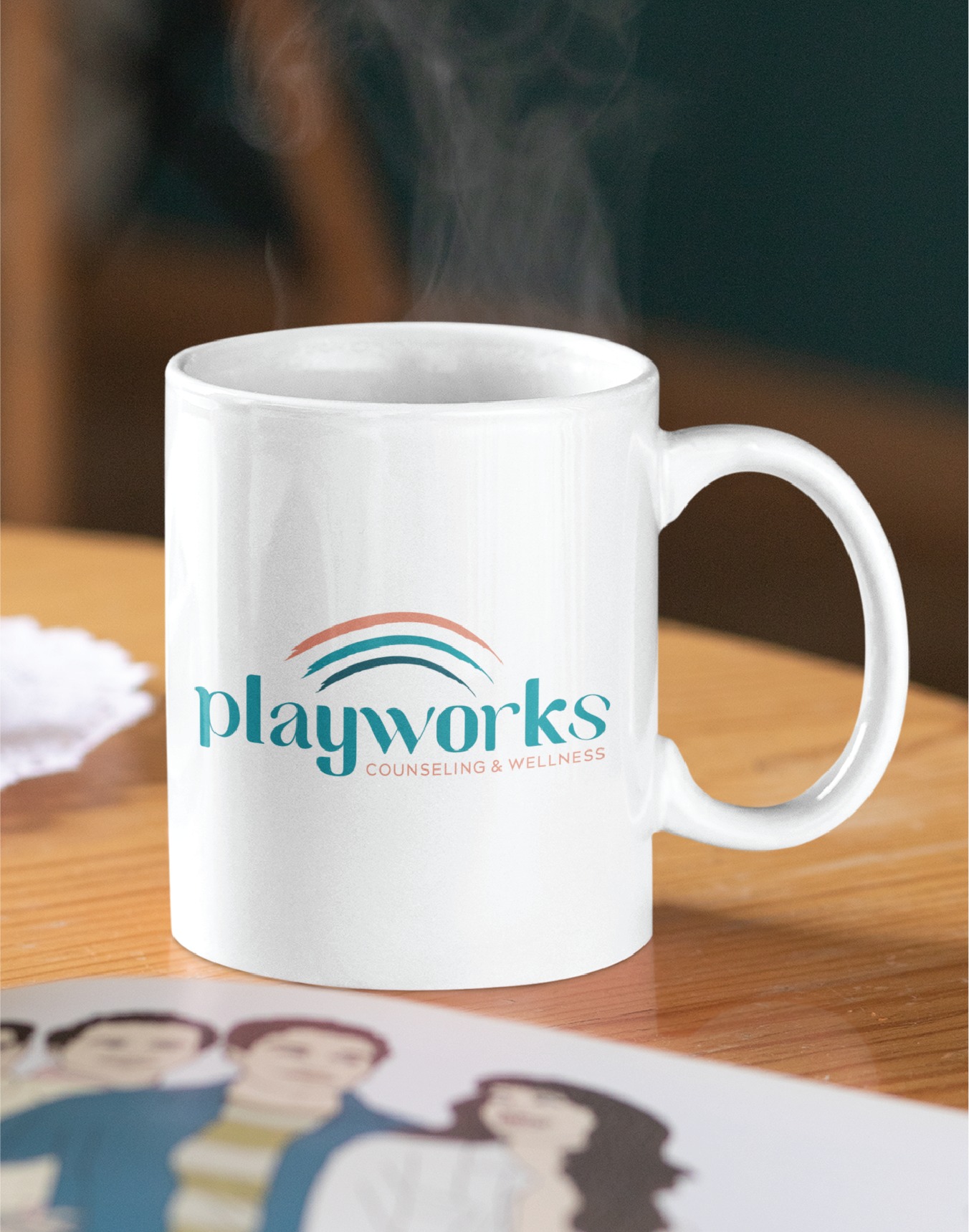 A white mug with the Playworks Counseling & Wellness custom logo design sits on a wooden table. Steam rises from the mug, and part of an illustrated image of people—a subtle nod to its roots in Virginia children's therapy—is partially visible in the foreground.