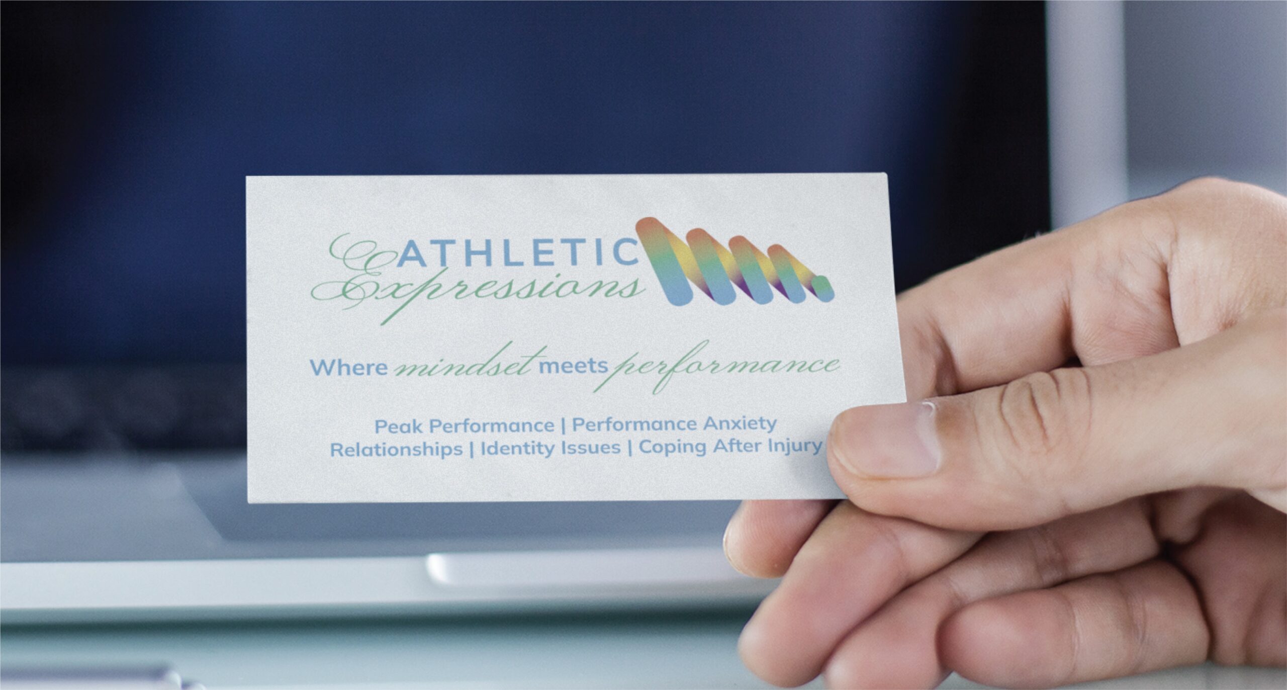 Business card reading'Athletic Expressions' with a colorful wave logo, held between fingers against a blurred background.