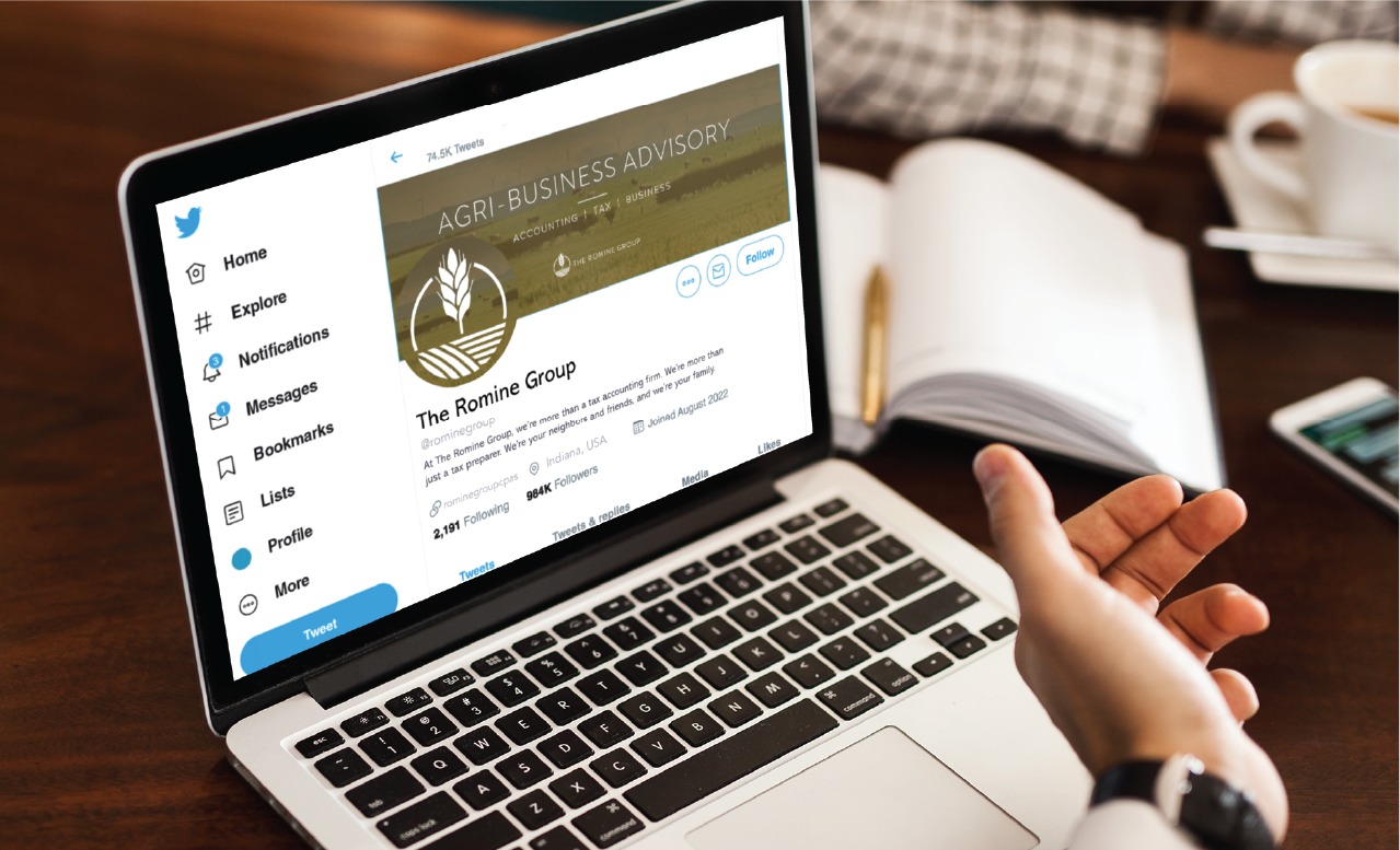 Laptop screen shows a Twitter profile for The Romine Group on a wooden desk with a hand in the foreground.