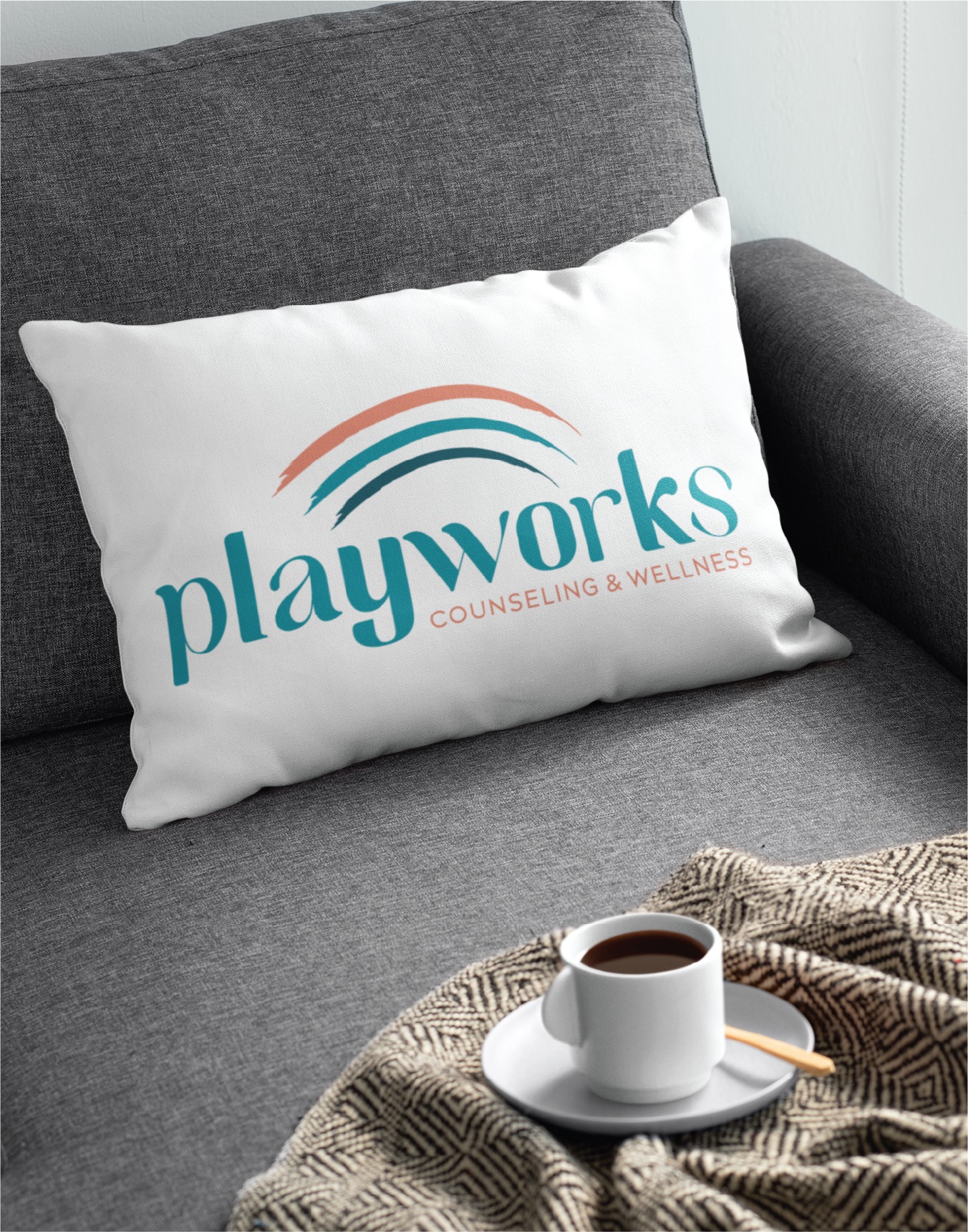 White pillow on a gray sofa featuring the Playworks Counseling & Wellness logo in teal and orange arcs.