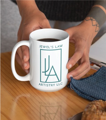 A person holding a white mug of coffee with the logo and text Jewel’s Law Artistry LLC—expert in Custom Logo Design and Jewelry Artist services in Virginia—printed in teal, next to a blue cloth and a pastry on a wooden table.