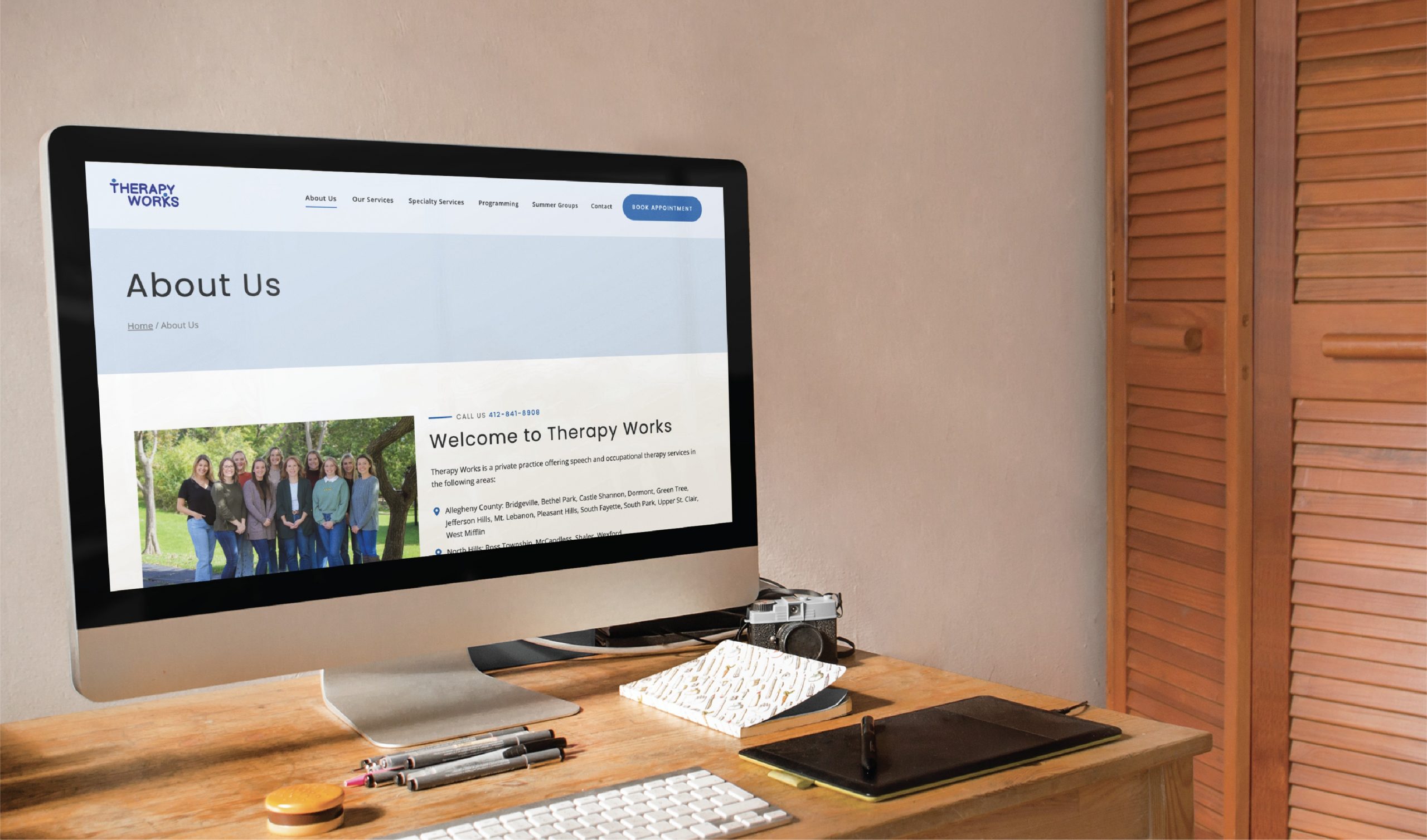A computer on a desk displays the “About Us” page for Therapy Works, highlighting their expertise in youth therapy and featuring a team photo alongside a welcome message. The desk is neatly arranged with a camera, notebook, eyeglasses, and pen. A louvered door sits in the background, creating a warm and inviting workspace that reflects Therapy Works’ commitment to Pennsylvania families through thoughtful website design.