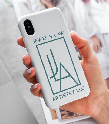 A hand holds a white phone case with the teal custom logo design and text: JEWELS LAW ARTISTRY LLC, proudly representing a talented Virginia jewelry artist. A magazine with blurred images is in the background.