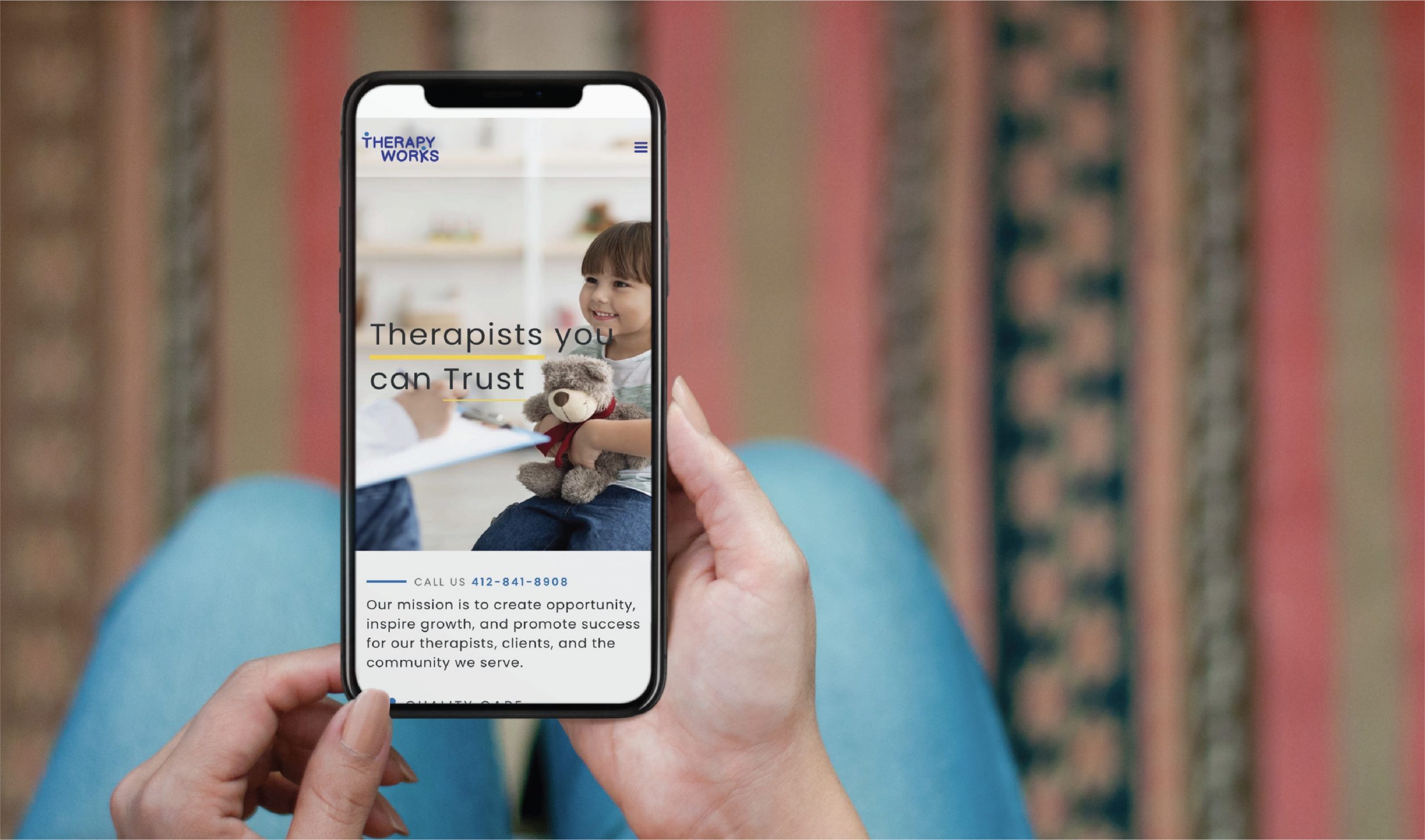 A person holds a phone displaying a Therapy Works website, featuring a smiling child holding a teddy bear and the text “Therapists you can trust.” Showcasing thoughtful website design tailored for youth therapy in Pennsylvania, the background reveals a patterned rug and blue jeans.