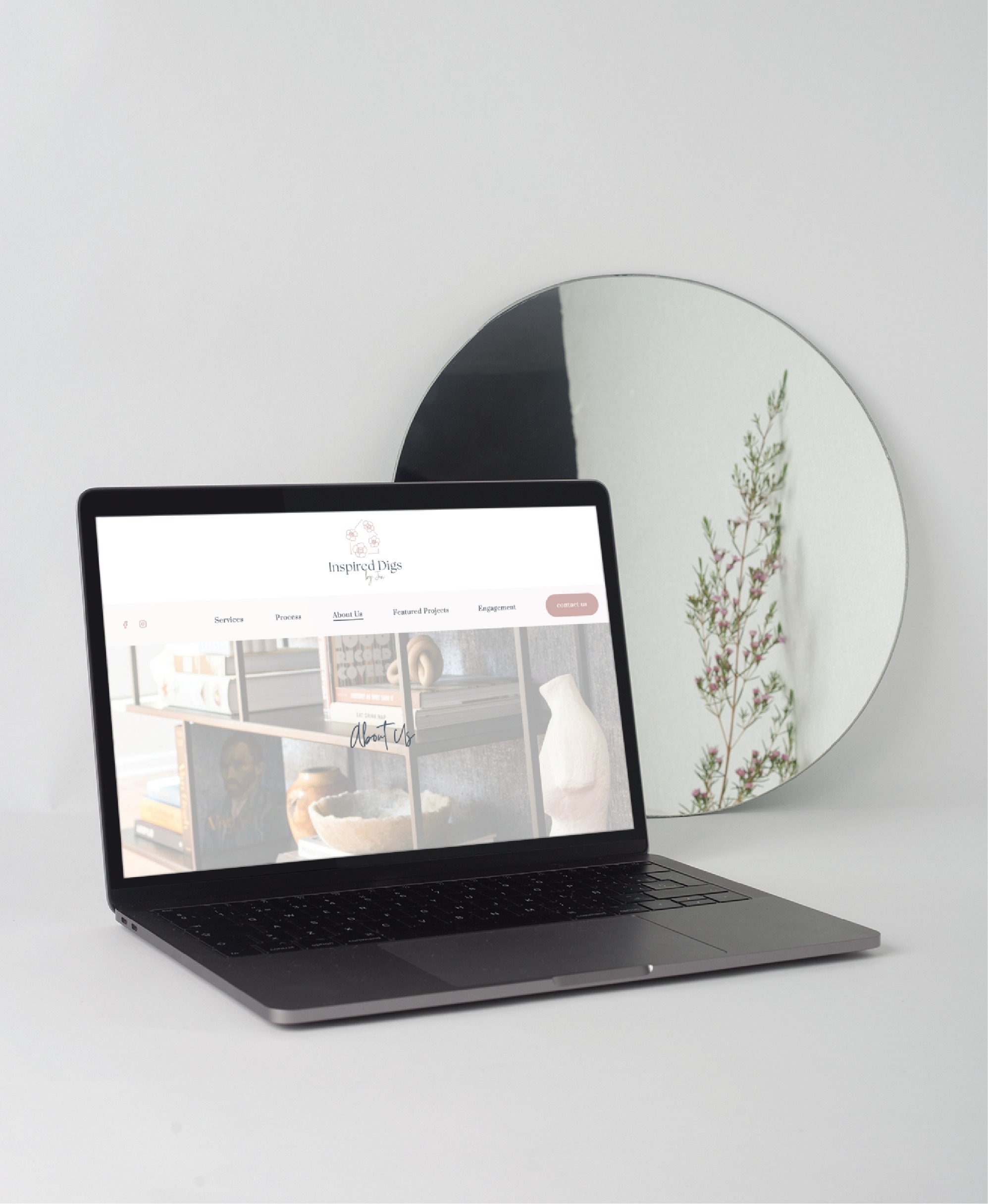 A laptop on a white surface displays a home decor website, showcasing elegant web design. Next to it, a round mirror reflects a sprig with small leaves against a light-colored wall.