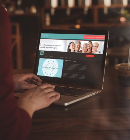 A person uses a laptop at a café in Pennsylvania. The screen shows a website with a banner image of smiling people and text, reflecting fresh social media design for a skincare brand. A cup of iced coffee sits on the table beside the laptop.