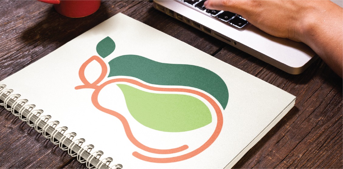 A hand uses a laptop beside a spiral notebook with a stylized, colorful pear illustration—ideal for logo creation—on the cover, placed on a wooden table next to a red coffee cup.