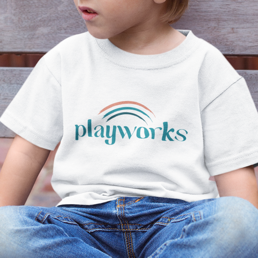 A young child sits on a bench wearing a white T-shirt with the word playworks and a stylized rainbow design, featuring a custom logo design often used by children’s therapists in Virginia, printed on the front.