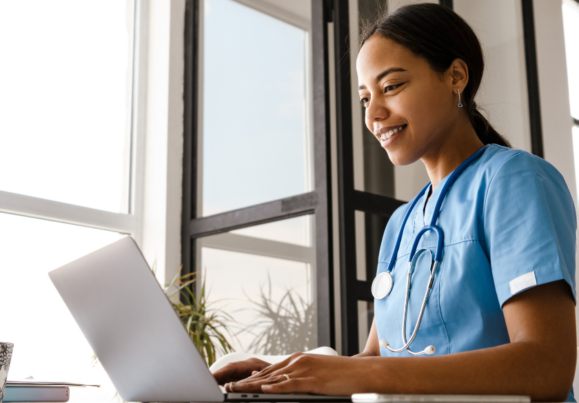 A smiling healthcare professional in blue scrubs with a stethoscope works on a laptop at a bright, modern desk near large windows, updating the new patient page on the medical practice website to help build a better experience for incoming patients.
