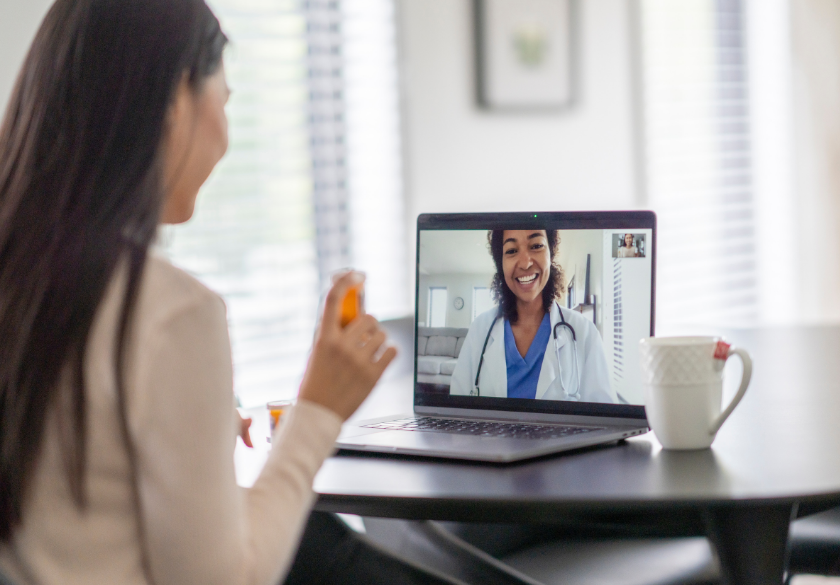 A woman holds a pill bottle while having a video call with a smiling doctor on her laptop at a table with a coffee mug—an example of how to build trust and convenience for patients when designing your medical practice website or new patient page.