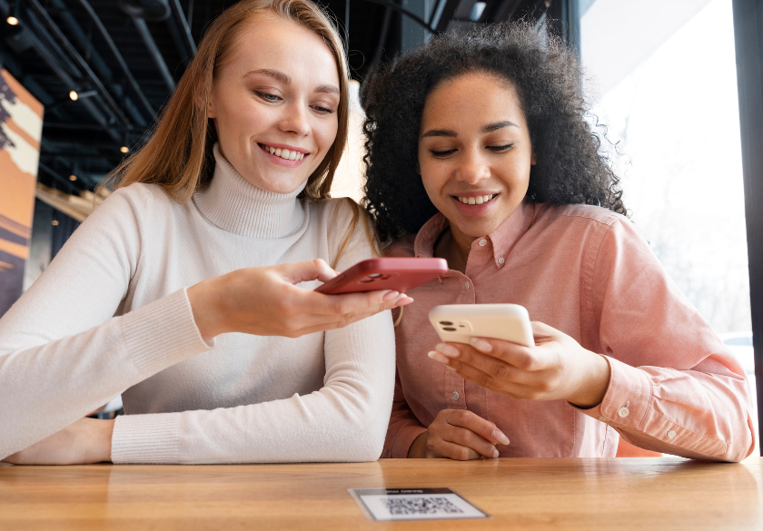 Two women smiling at their smartphones while sitting at a wooden table, a QR code visible on the table in front of them in a bright cafe.