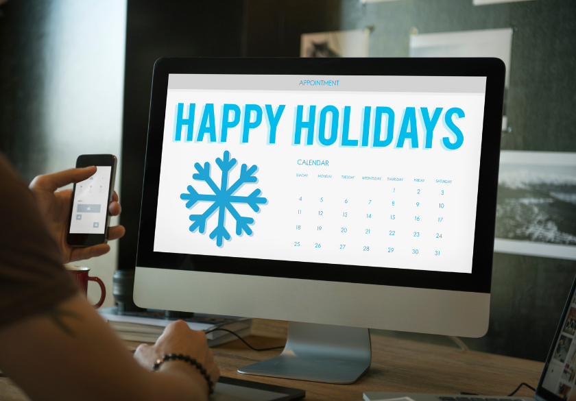 A computer screen displays HAPPY HOLIDAYS with a blue snowflake and a calendar, capturing the festive spirit of Q4. A person holds a smartphone and sits at a desk with a keyboard, notebook, and pen—perfectly poised to plan some effective holiday marketing for their small business website.