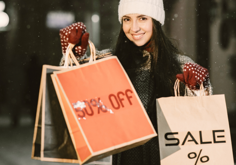 A woman in winter clothes smiles while holding shopping bags, one labeled 50% OFF and another labeled SALE %, capturing the excitement of holiday marketing in Q4. Snow is falling in the background, evoking the festive spirit that small business websites aim to inspire during this busy shopping season.
