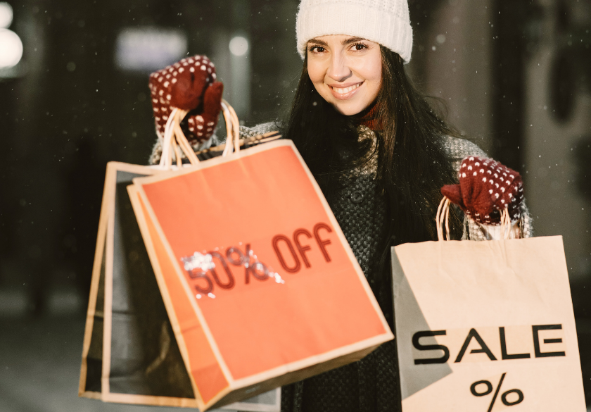 A woman in winter clothes smiles while holding shopping bags, one labeled 50% OFF and another labeled SALE %, capturing the excitement of holiday marketing in Q4. Snow is falling in the background, evoking the festive spirit that small business websites aim to inspire during this busy shopping season.