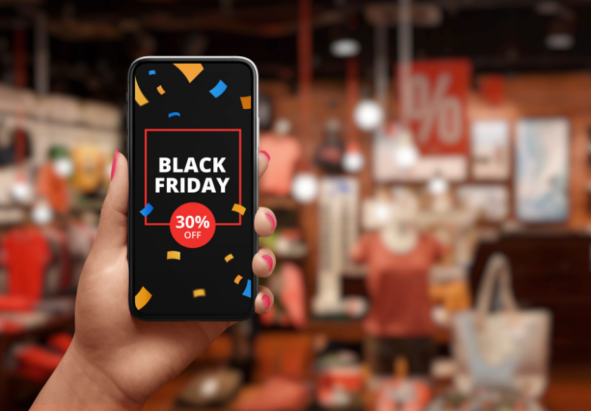 A hand holds a smartphone displaying a Black Friday 30% OFF sign with confetti, showcasing an exciting example of holiday marketing against a blurred background of a clothing store.