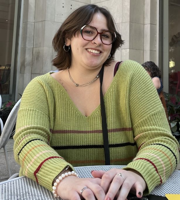 A person with short brown hair and glasses, wearing a green sweater, smiles while sitting at an outdoor cafe table, discussing web design projects for her woman-owned business.