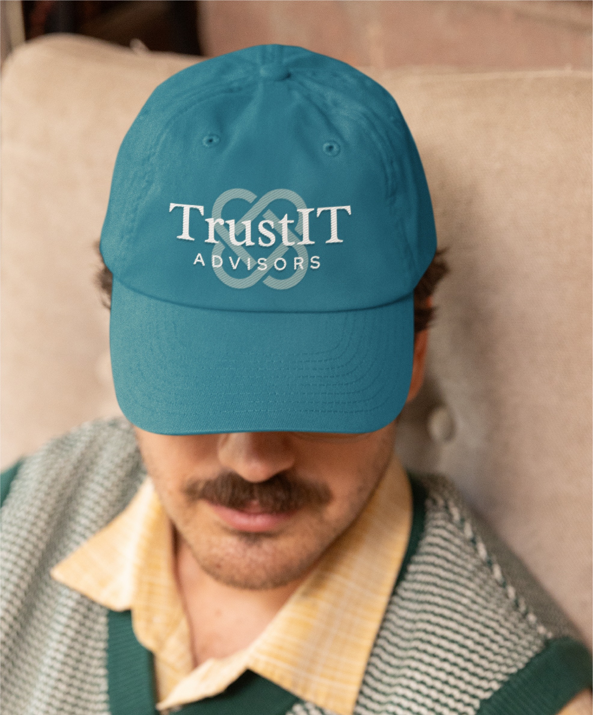 A person with a mustache wears a teal baseball cap that reads TrustIT ADVISORS in white letters, showcasing a professional logo design. The person, likely one of the skilled IT consultants from Texas, is sitting indoors on a light-colored chair, wearing a yellow shirt and green vest.