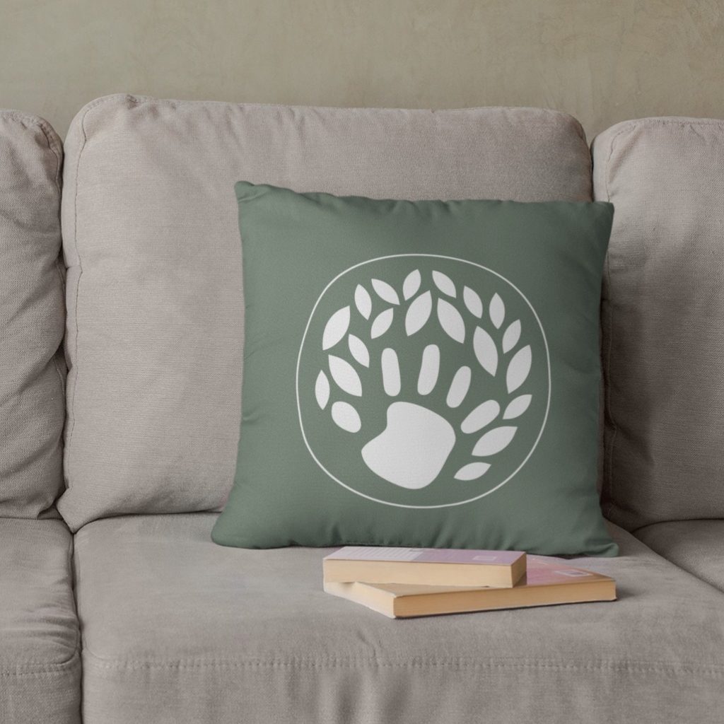 A green throw pillow with a white design featuring a handprint and leaf shapes—reminiscent of a children’s therapist logo design—rests on a light gray couch. Two closed books with pastel covers lie in front of the pillow, adding a cozy touch to this inviting Virginia home setting.