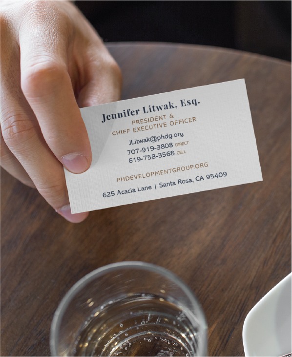 A hand holding a business card featuring elegant stationery design for Jennifer Litwak, Esq., President and CEO, with contact info, rests above a glass of water on a wooden table.