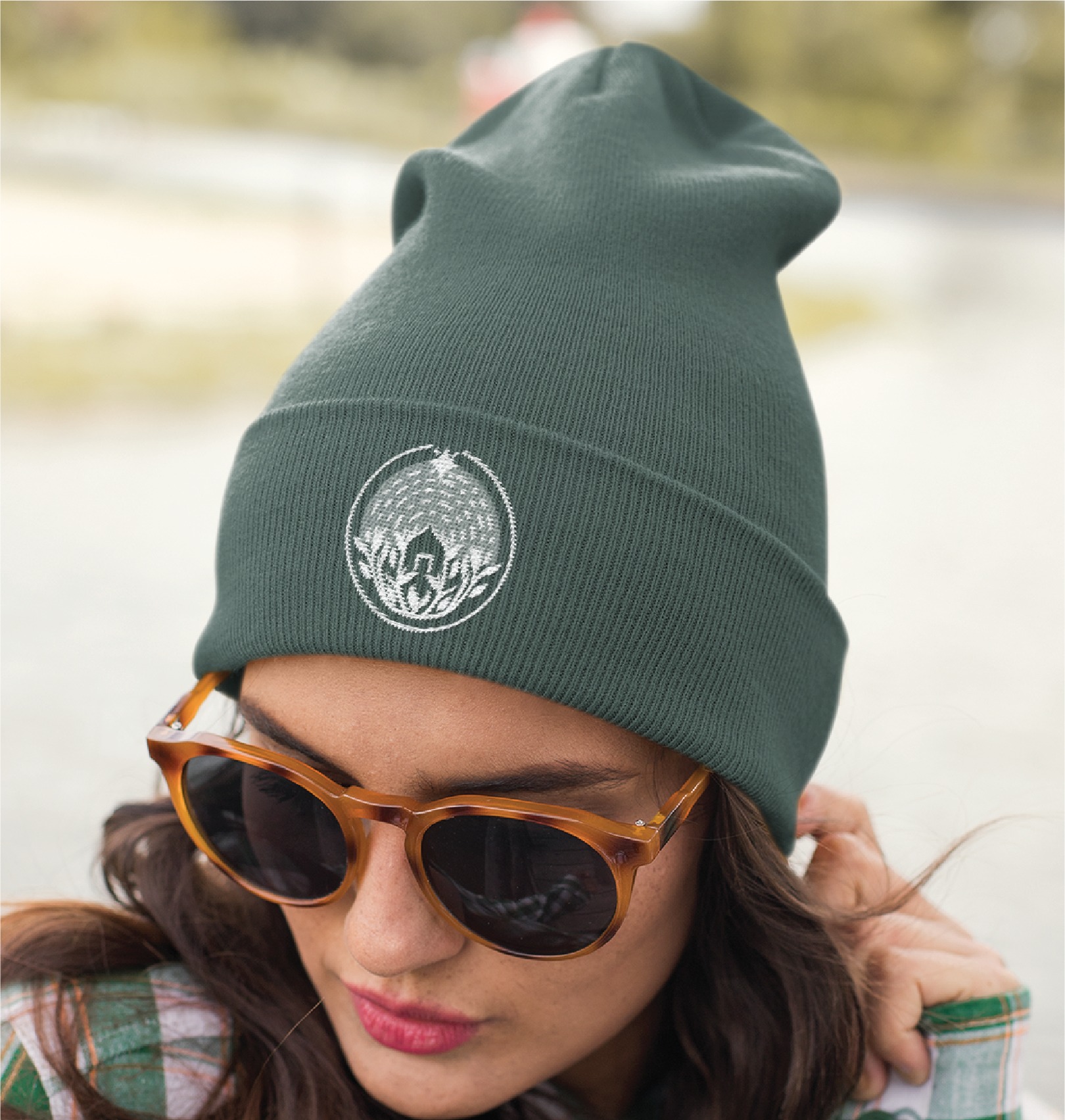 Woman wearing brown sunglasses and a dark green beanie with a white embroidered logo illustration, looking downward. The background is blurred, suggesting an outdoor California setting.