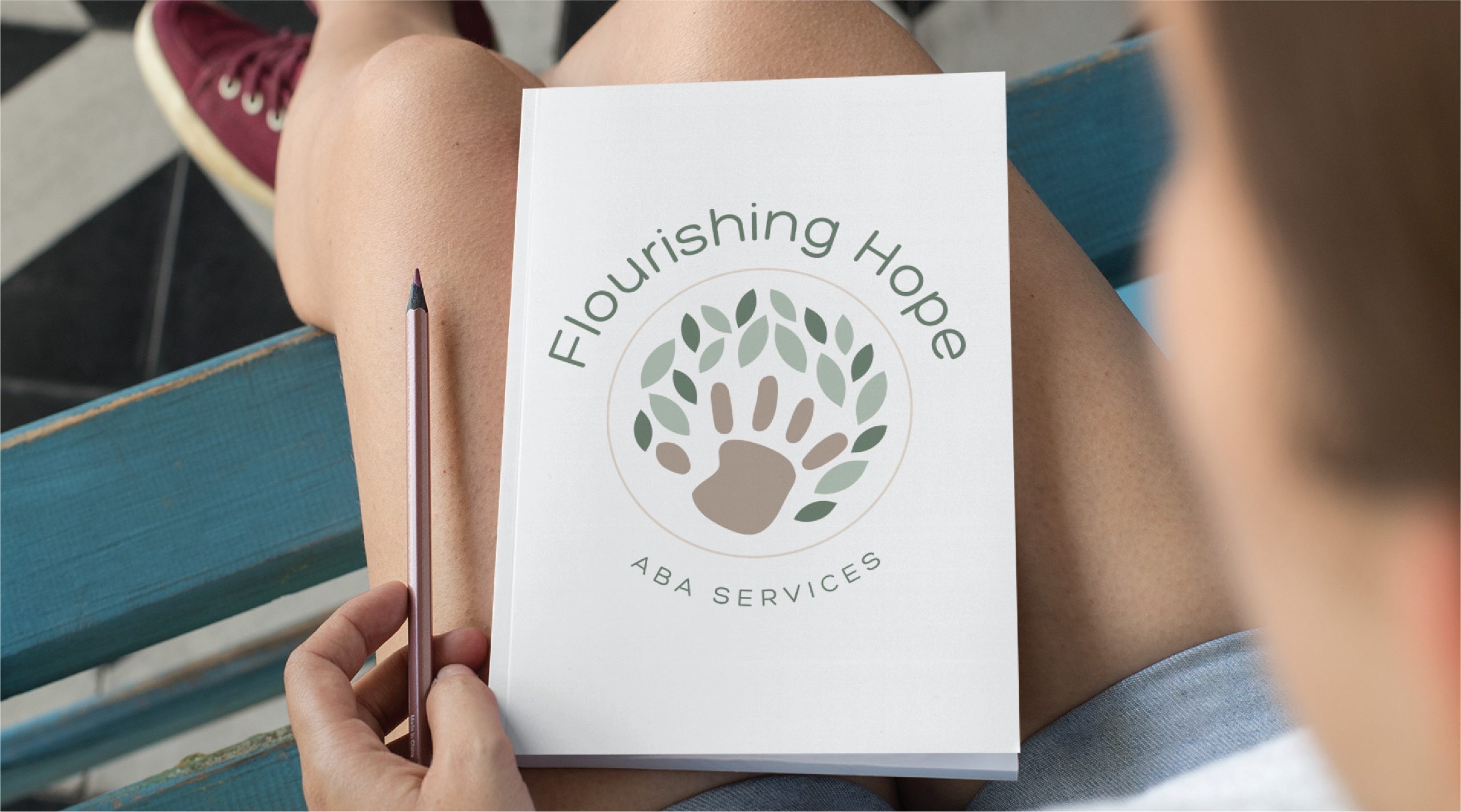 A person holds a notebook with the cover reading Flourishing Hope ABA Services and a logo of a handprint surrounded by green leaves, reflecting thoughtful logo design for a children’s therapist in Virginia. The person sits on a blue bench and holds a pencil.
