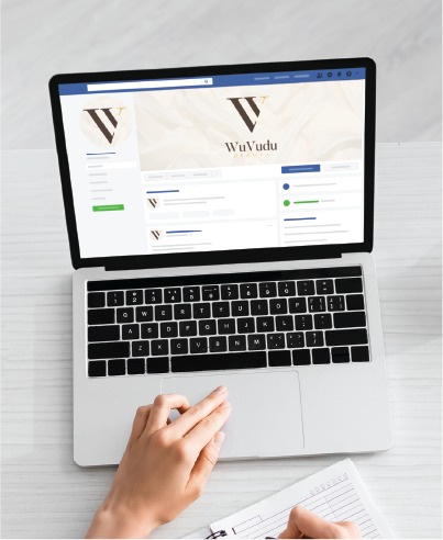 A person uses a laptop displaying a WuVudu social media profile page featuring innovative Social Media Design while writing in a notebook on a white desk.
