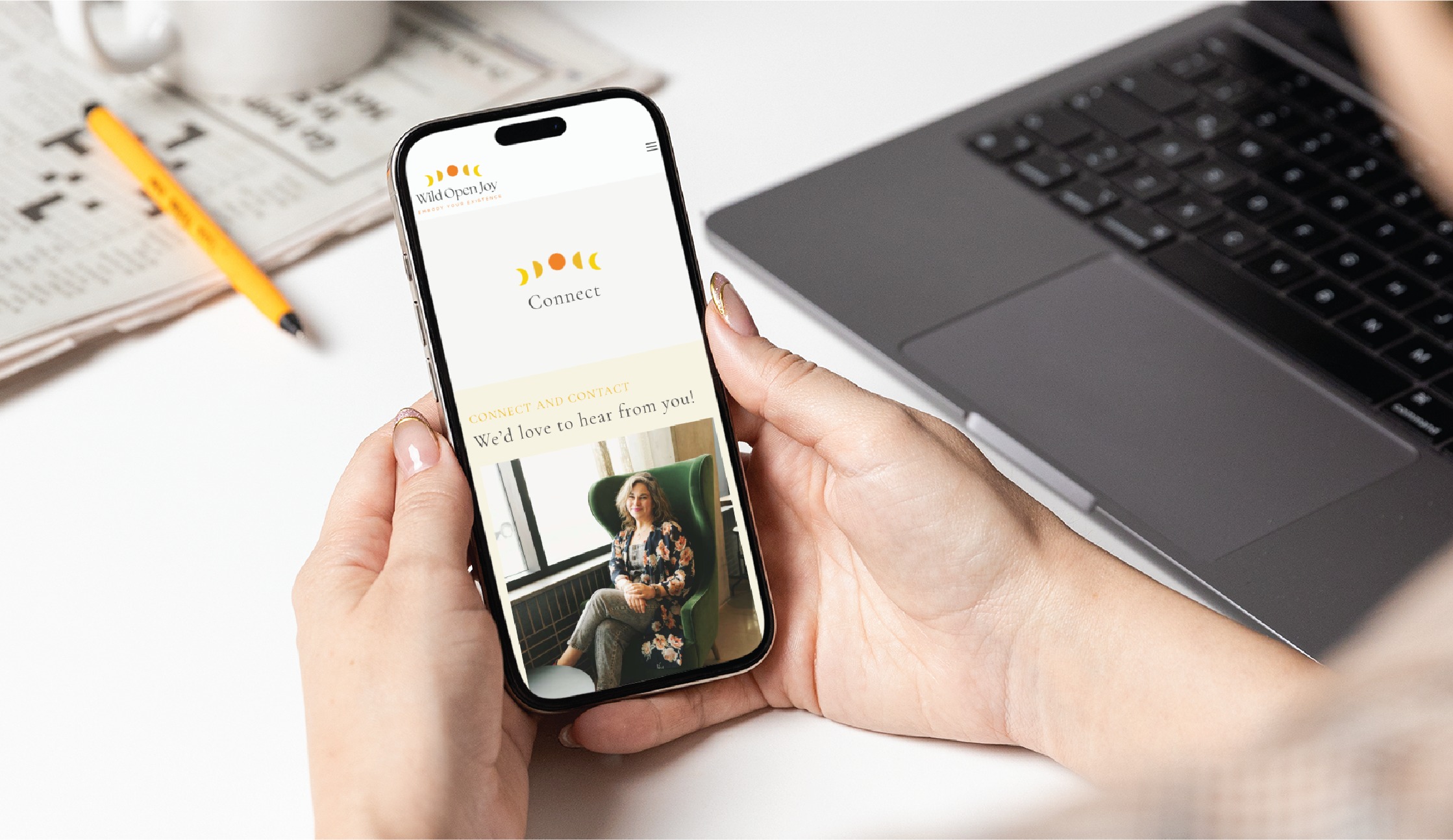 A person holds a smartphone displaying a website contact page with a woman’s photo, highlighting thoughtful website design. A laptop, pencil, coffee cup, and newspaper are visible on the desk in the background, creating a productive workspace atmosphere.