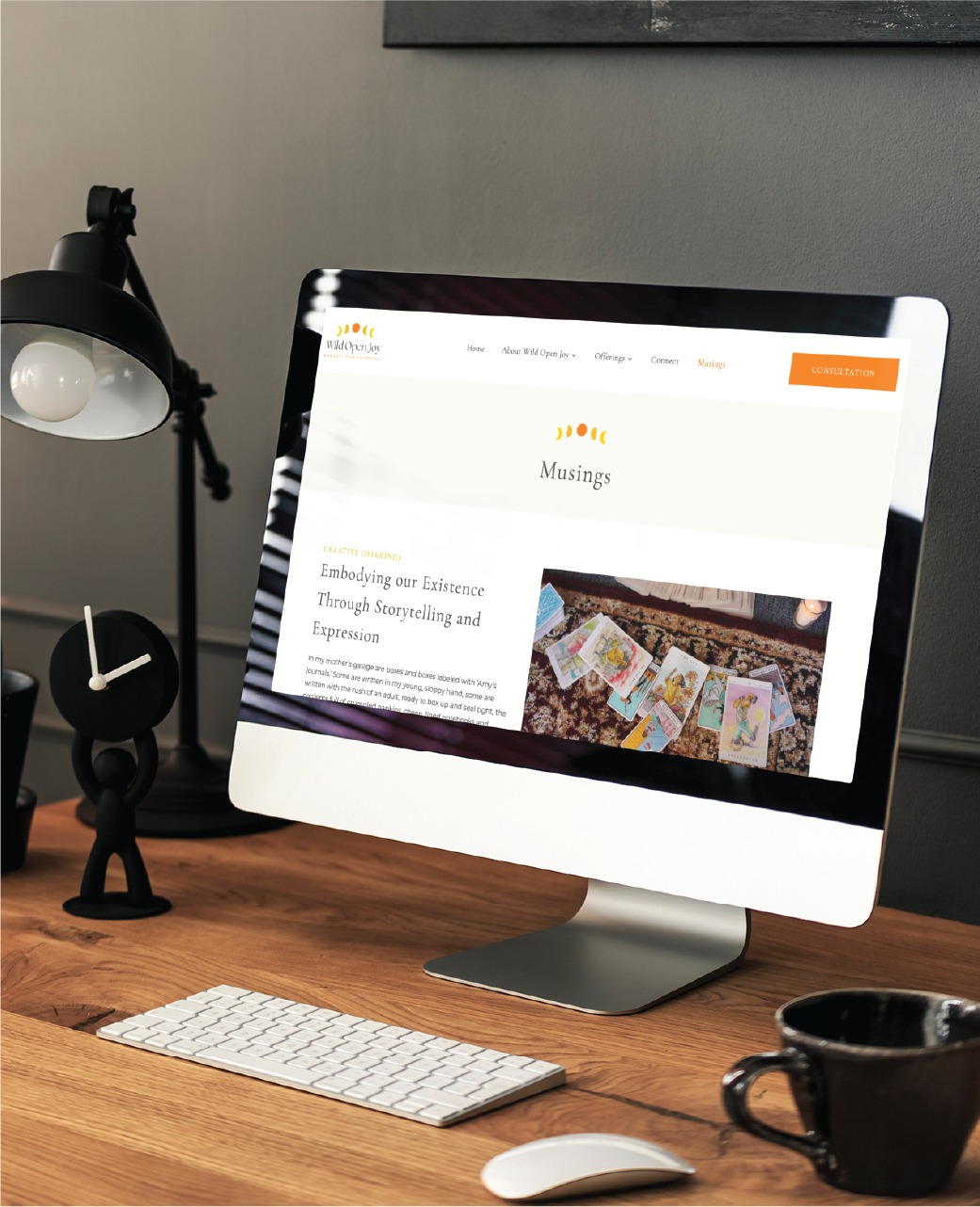 A computer on a wooden desk displays a blog webpage titled Musings with tarot cards pictured, reflecting the thoughtful perspective of a Spiritual Therapist in Colorado. Nearby are a lamp, a clock, a keyboard, a mouse, and a small white dish—details that complement the inviting website design.