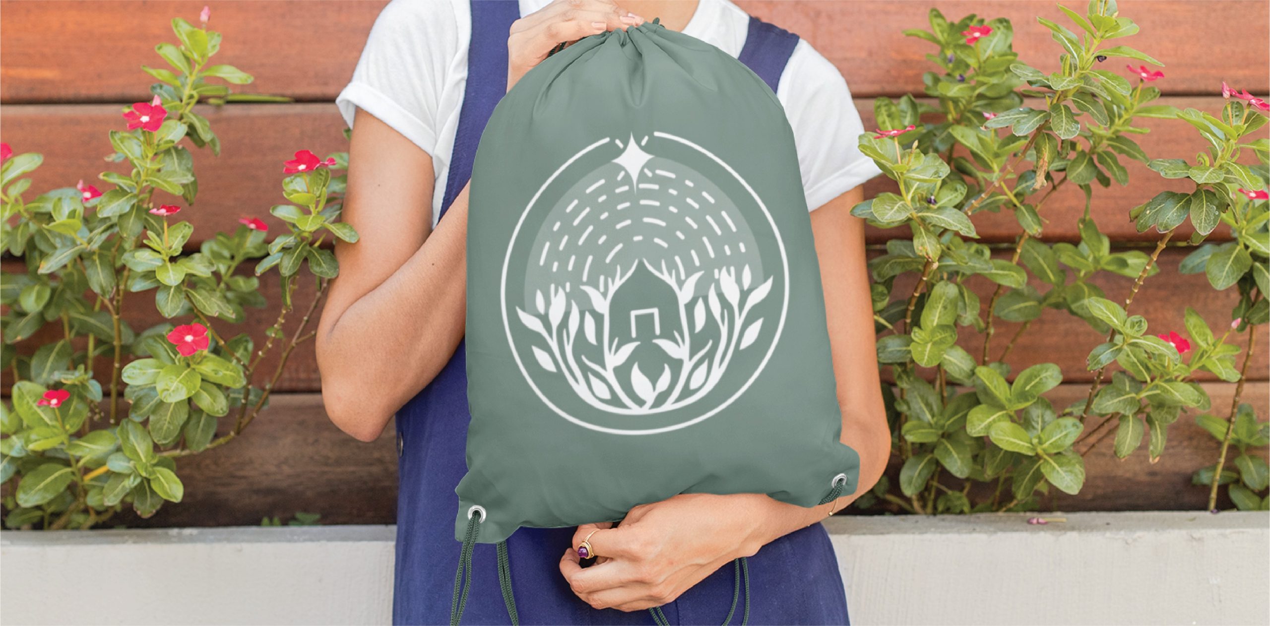 A person in a blue apron holds a green drawstring bag featuring a white nature-inspired logo illustration, standing in front of a planter blooming with pink flowers and lush green leaves—a perfect scene for California garden care enthusiasts.