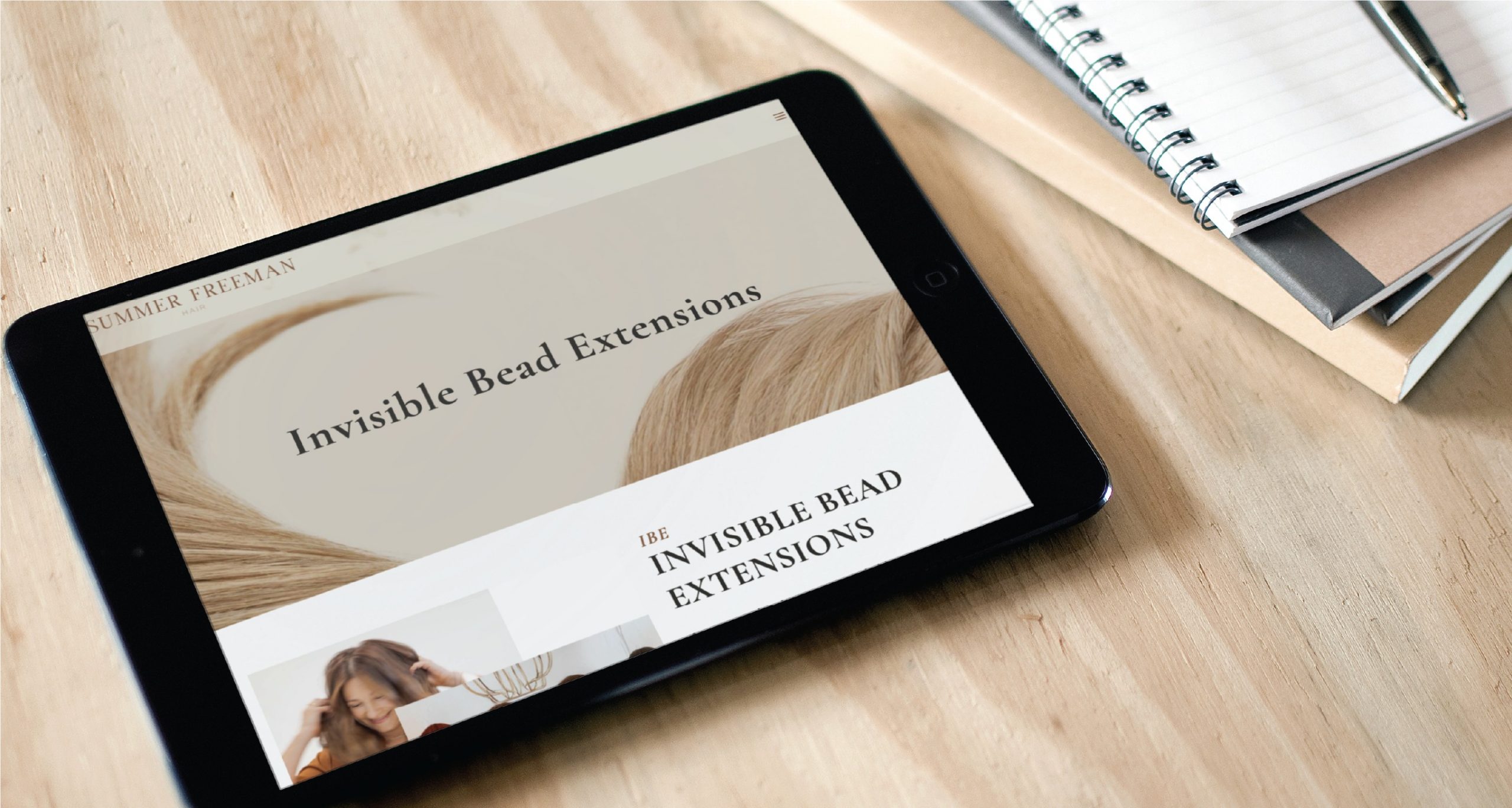 A tablet on a wooden desk displays a website about invisible bead hair extensions, showcasing sleek website design ideal for any California hairdresser. Next to it are a spiral notebook, a pen, and a closed notebook.