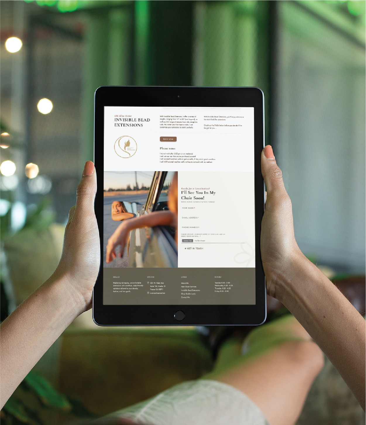 A person holds a tablet displaying a salon website about invisible bead extensions, designed with elegant website design elements. The site features a logo, text, a photo of a woman on a boat, and contact details for the California-based hairdresser. The background shows a blurred indoor setting with plants.