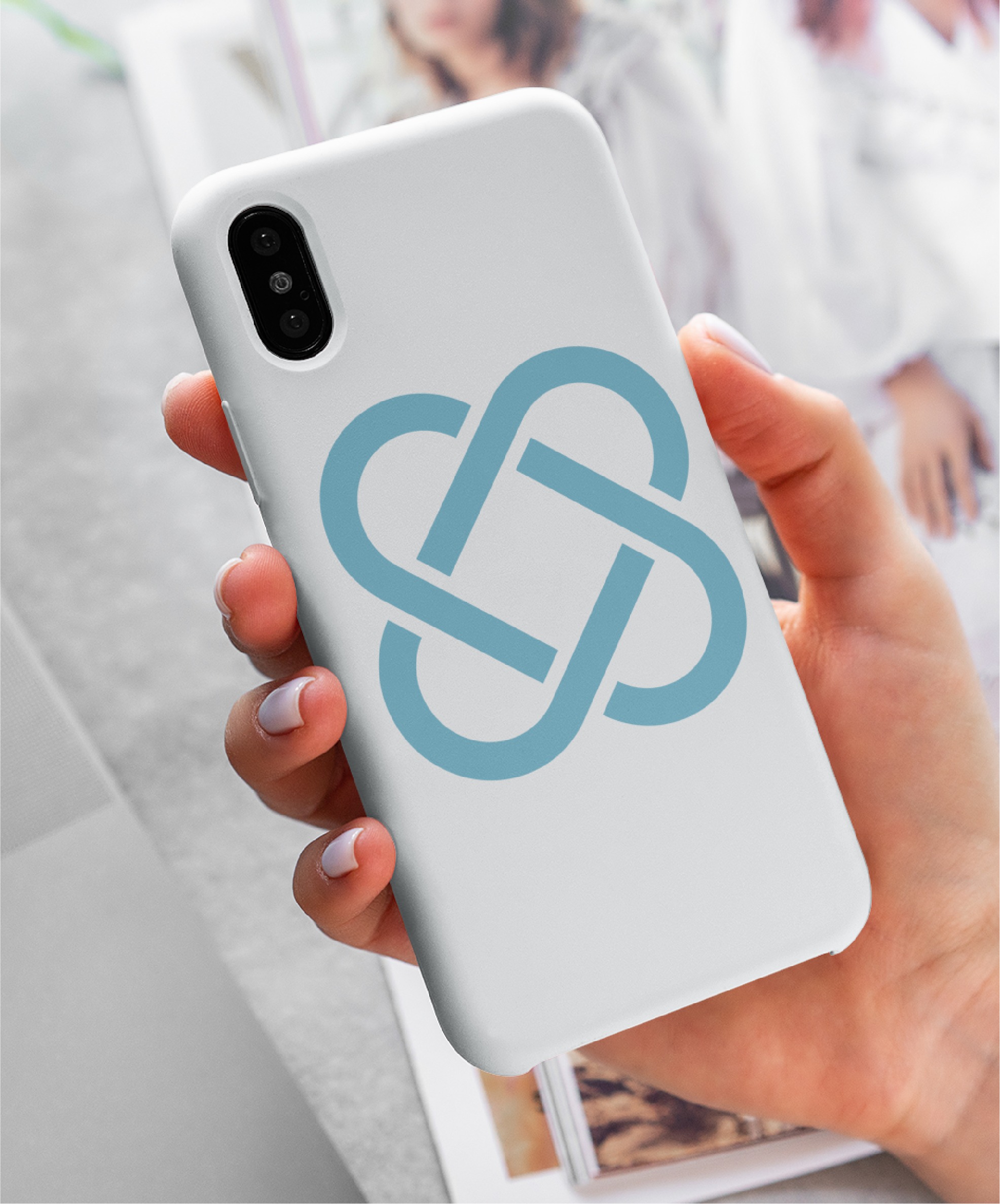 A hand holds a white smartphone with a blue geometric logo, reflecting expert logo design, on the case. In the background, there are blurred magazines with images of people—hinting at dynamic Texas IT consultants and creative innovation.