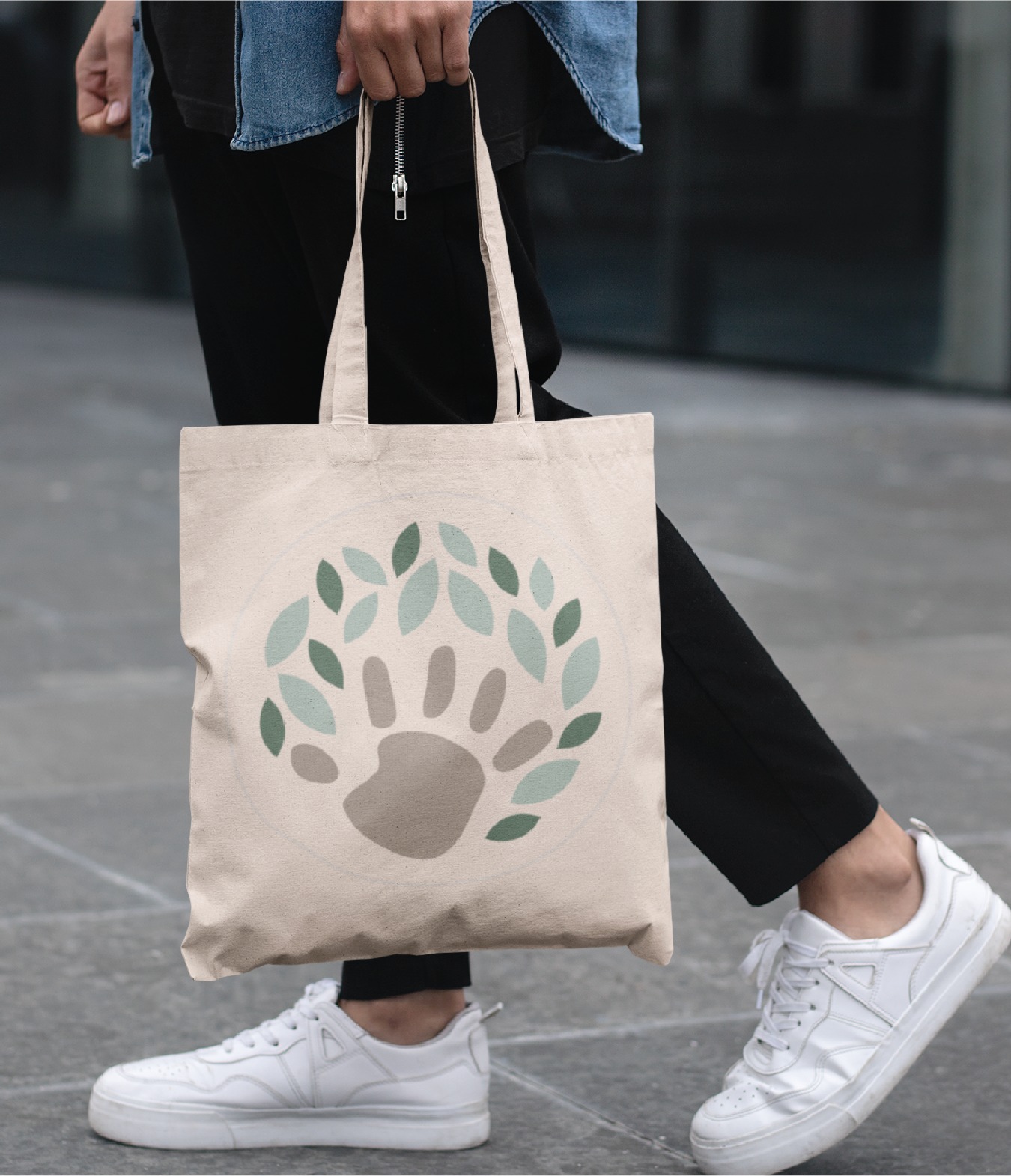 A person walking on a gray sidewalk in Virginia holds a beige tote bag featuring a logo design of a paw print surrounded by green leaves. They wear white sneakers, black pants, and a blue top.