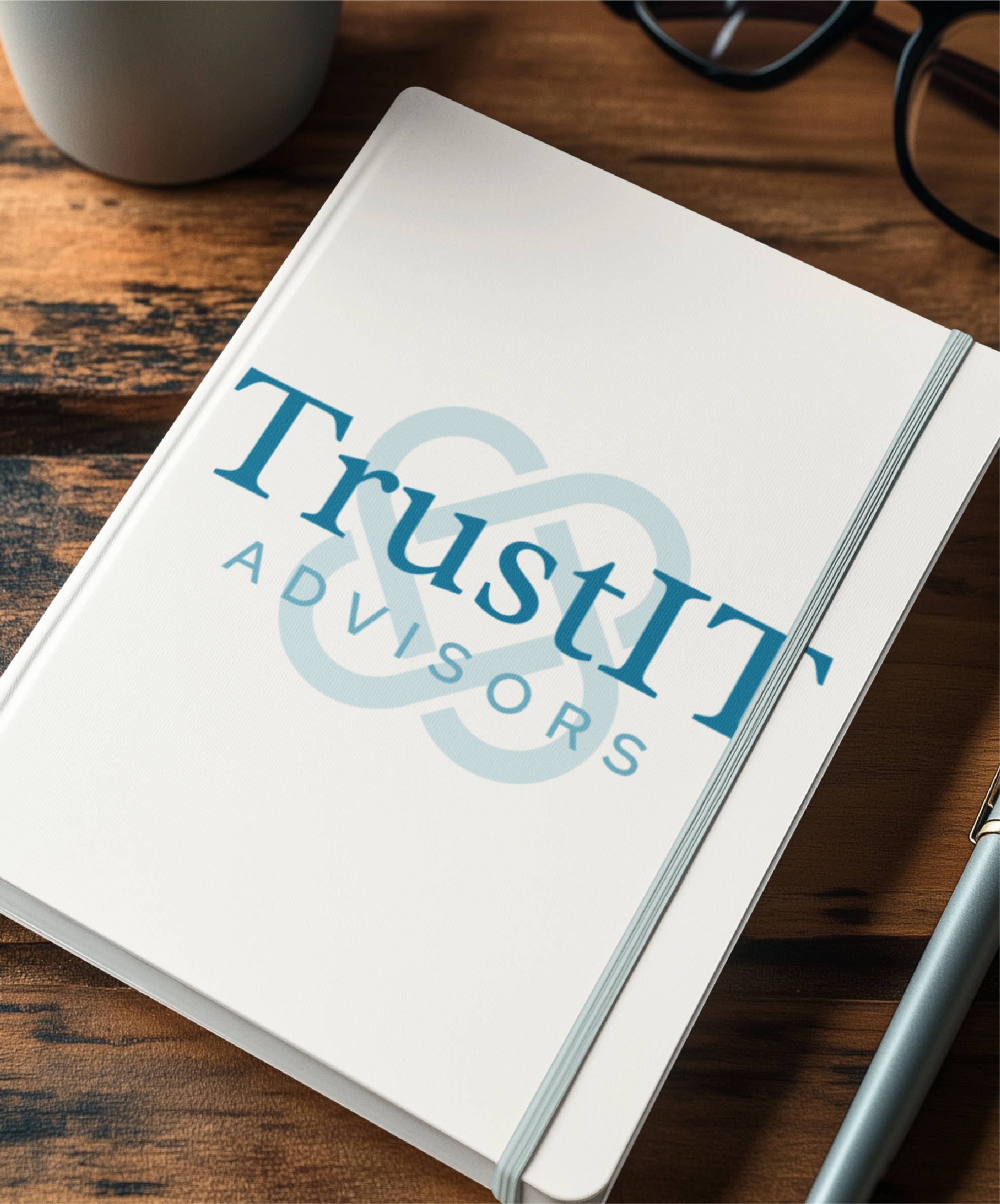 A white notebook featuring the TrustIT Advisors logo design on the cover lies on a wooden desk next to a pen, glasses, and a coffee cup—the perfect desktop setup for Texas-based IT consultants.