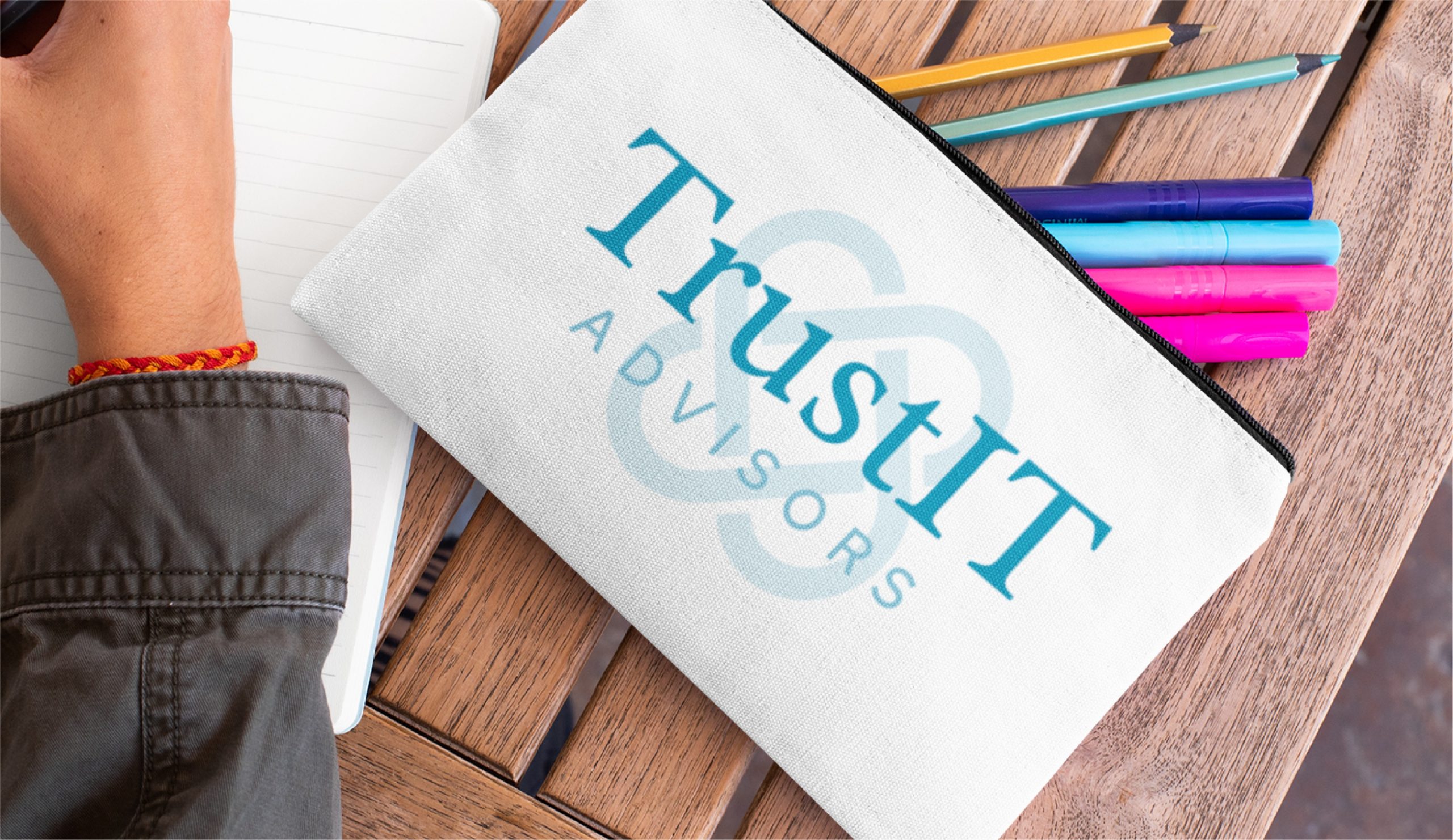 A hand writes in a notebook next to a zippered pouch labeled TrustIT Advisors, surrounded by colorful pens and pencils on a wooden table—a scene that captures the creativity and organization trusted by Texas IT Consultants and logo design professionals alike.